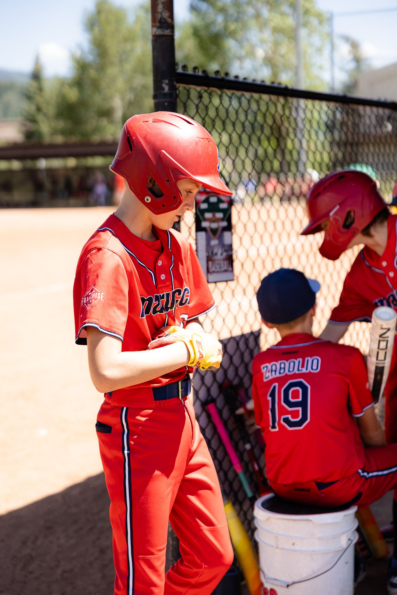 Young baseball player in red uniform and helmet adjusts gloves near dugout fence during a youth baseball game.