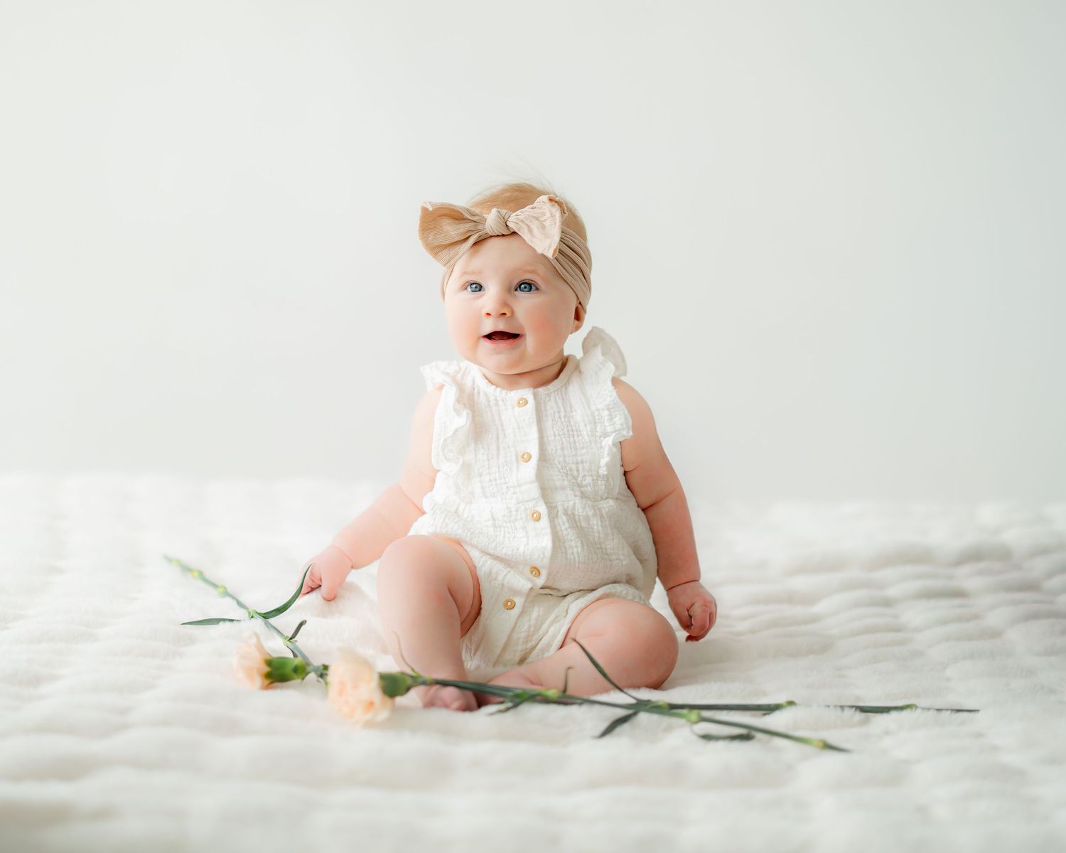 A baby in a white dress sits on a white blanket with a single pink flower.