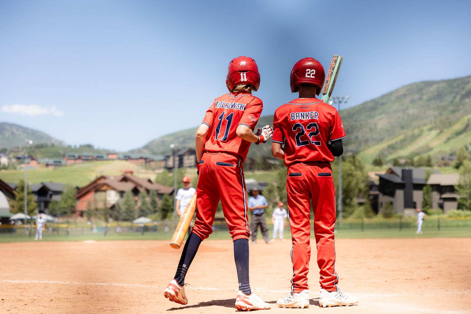 Two youth baseball players in red uniforms standing together on dirt field with mountains behind.