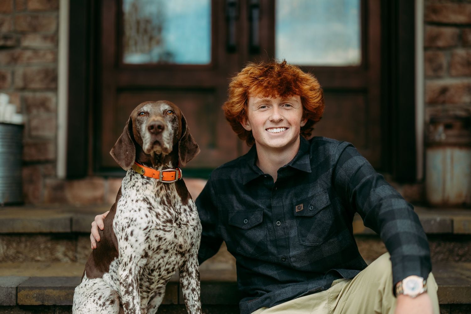 Person in dark clothing sitting with spotted dog against brick wall.