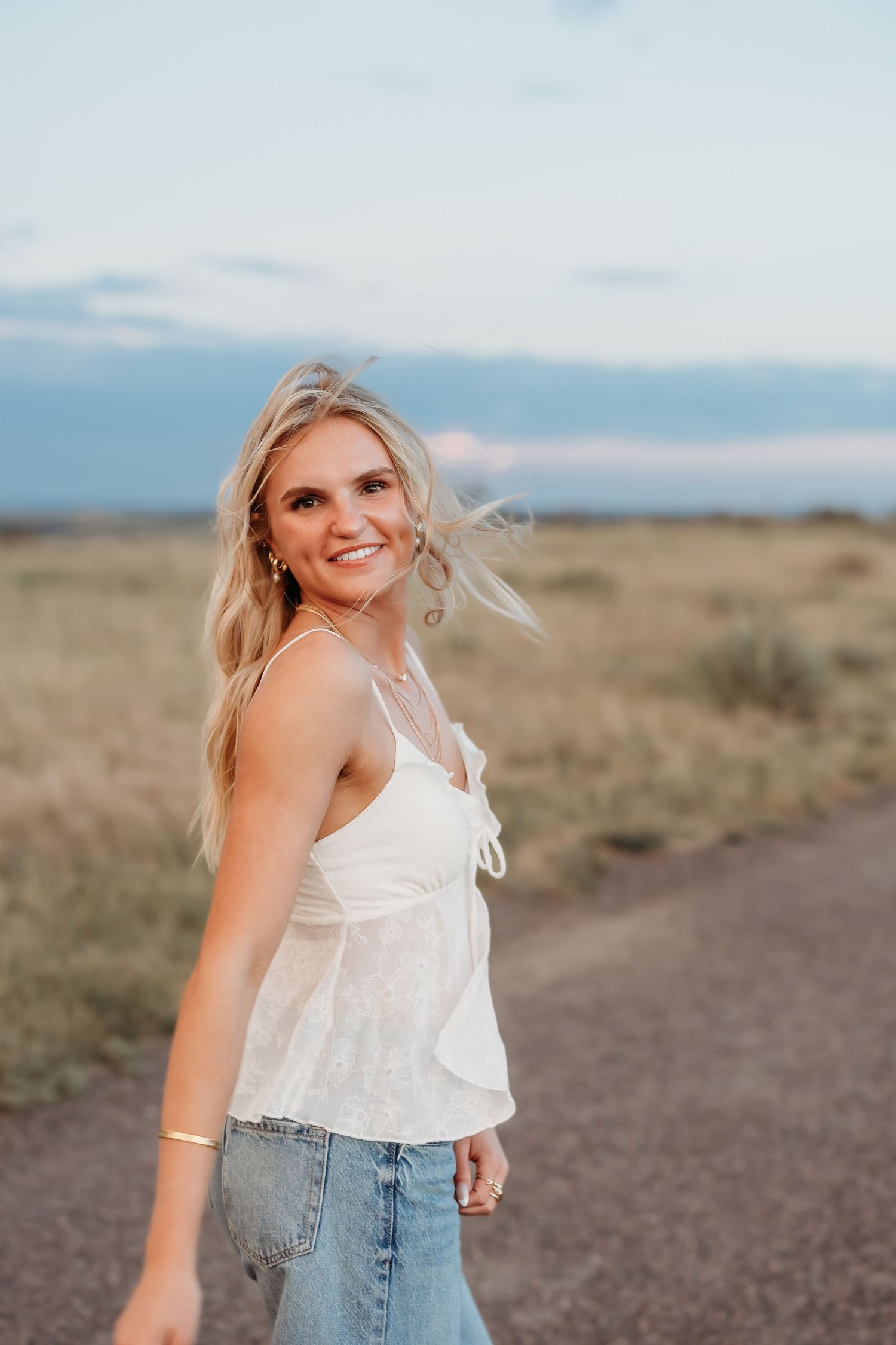 A person in a white tank top and jeans smiles while standing on a dirt path at sunset with wind-blown hair.