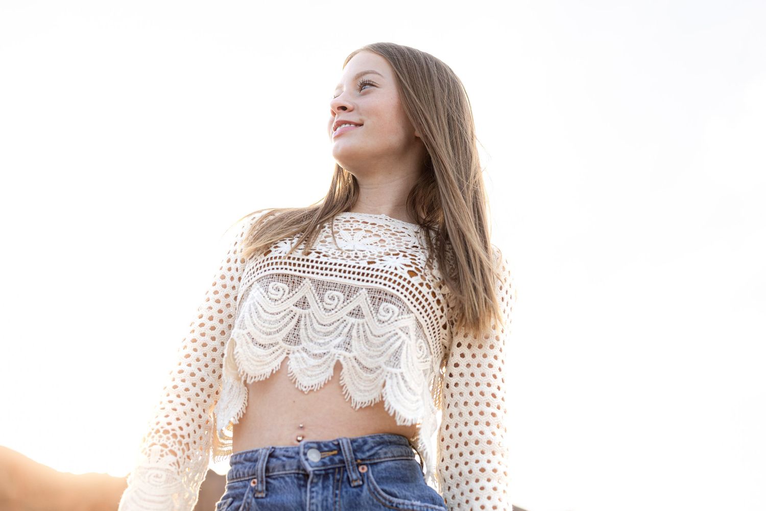 Woman wearing white lace top and jeans poses against bright white background.
