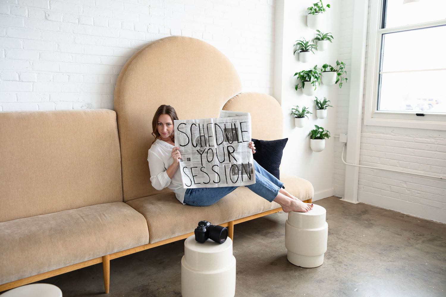 Woman sitting on beige sofa holding a handwritten sign reading Schedule Your Session in a modern studio.