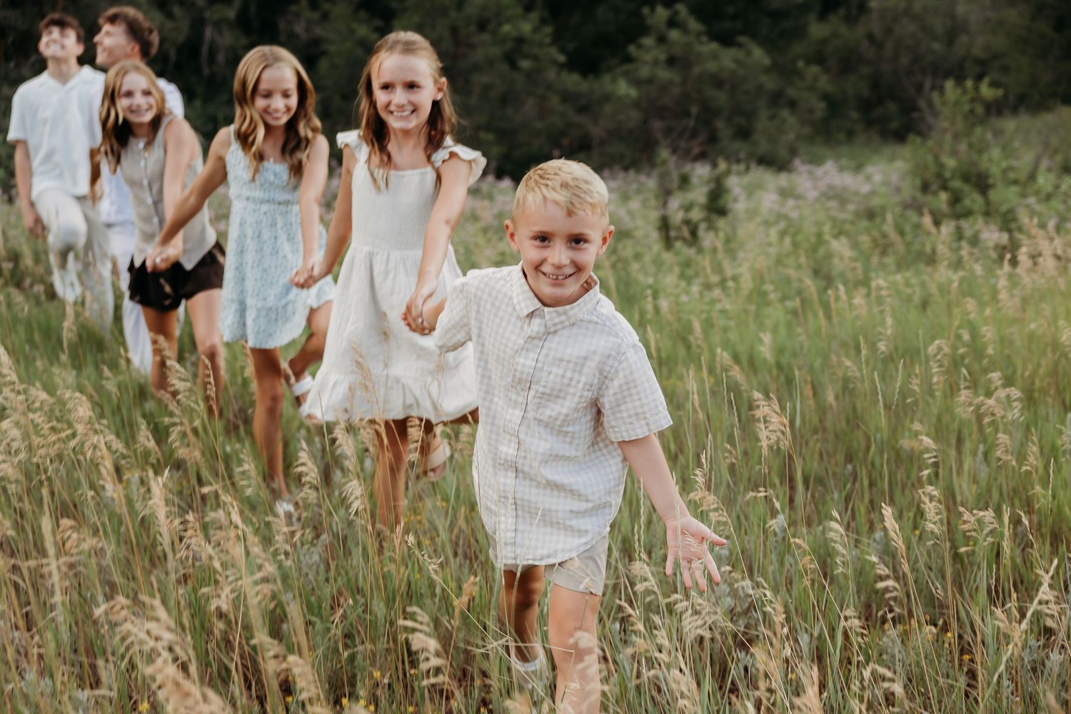 A family in white outfits walks through a sunlit meadow with tall grass during a summer photo session.