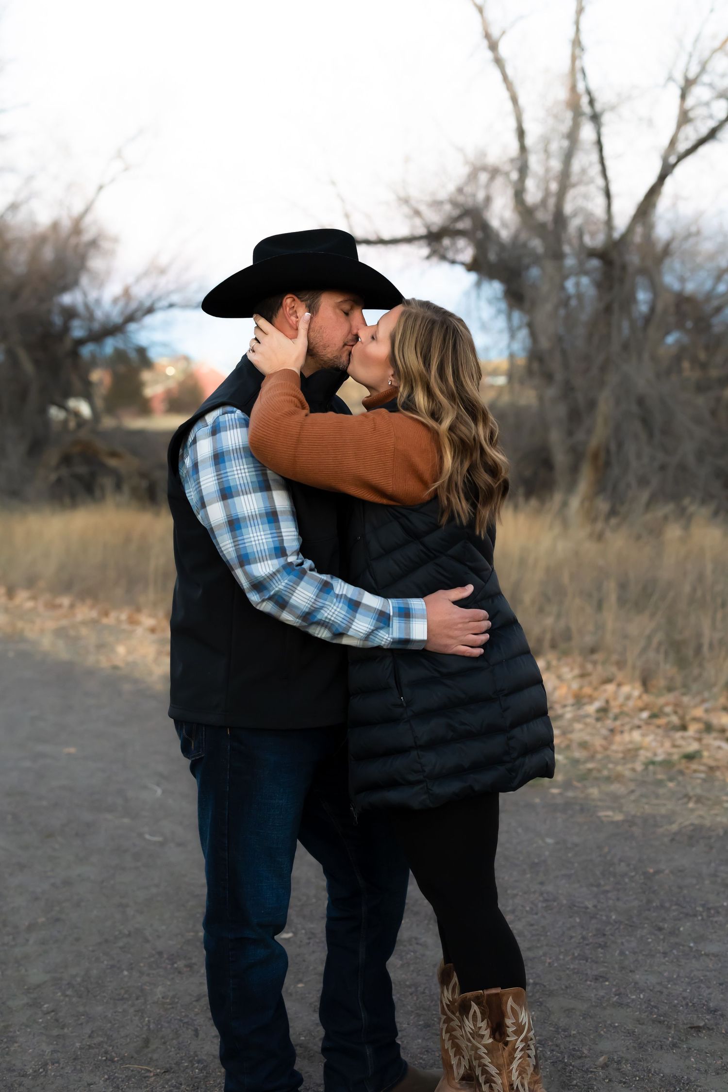 A couple embraces in a romantic outdoor setting wearing western attire and plaid shirt against a natural backdrop.