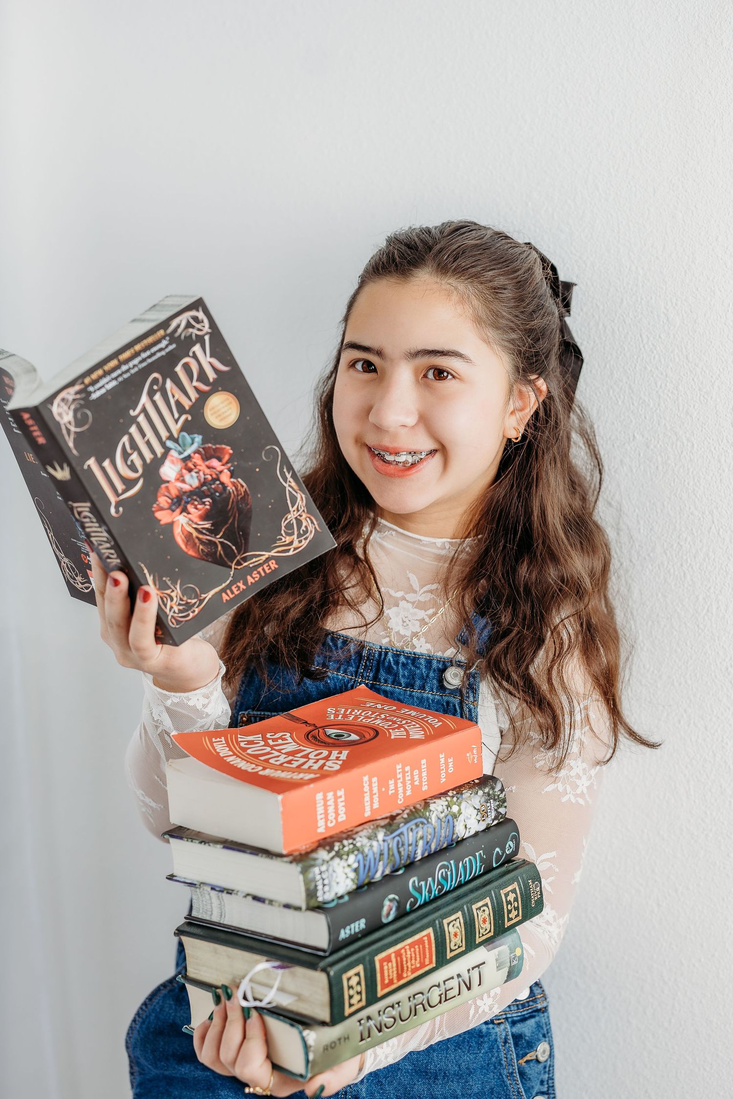 8th grade girl in denim overalls holds a stack of books including 'Lethal Omen' against a white wall.