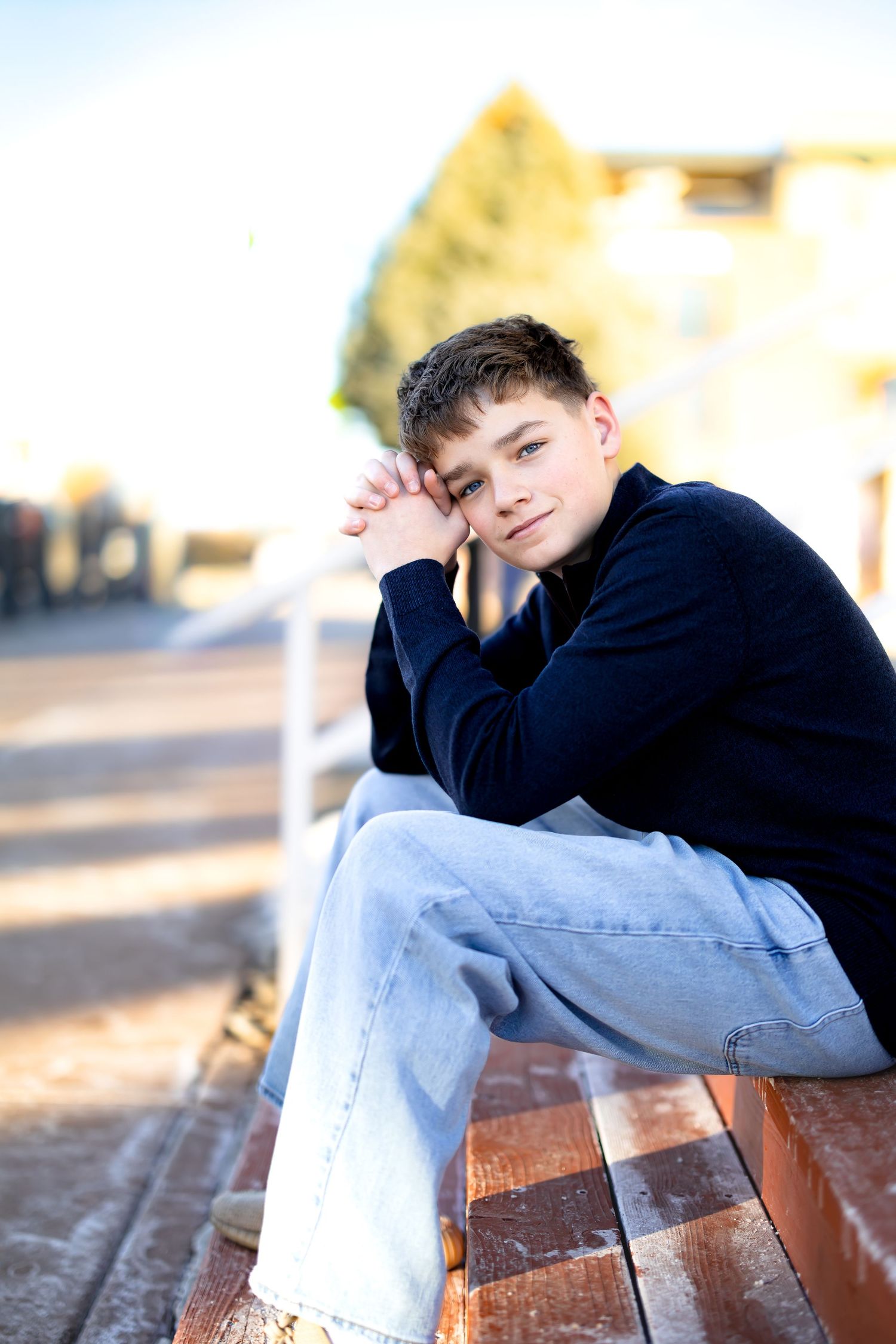 8th grade boy in a black sweater and jeans sits thoughtfully on wooden steps in warm outdoor lighting.