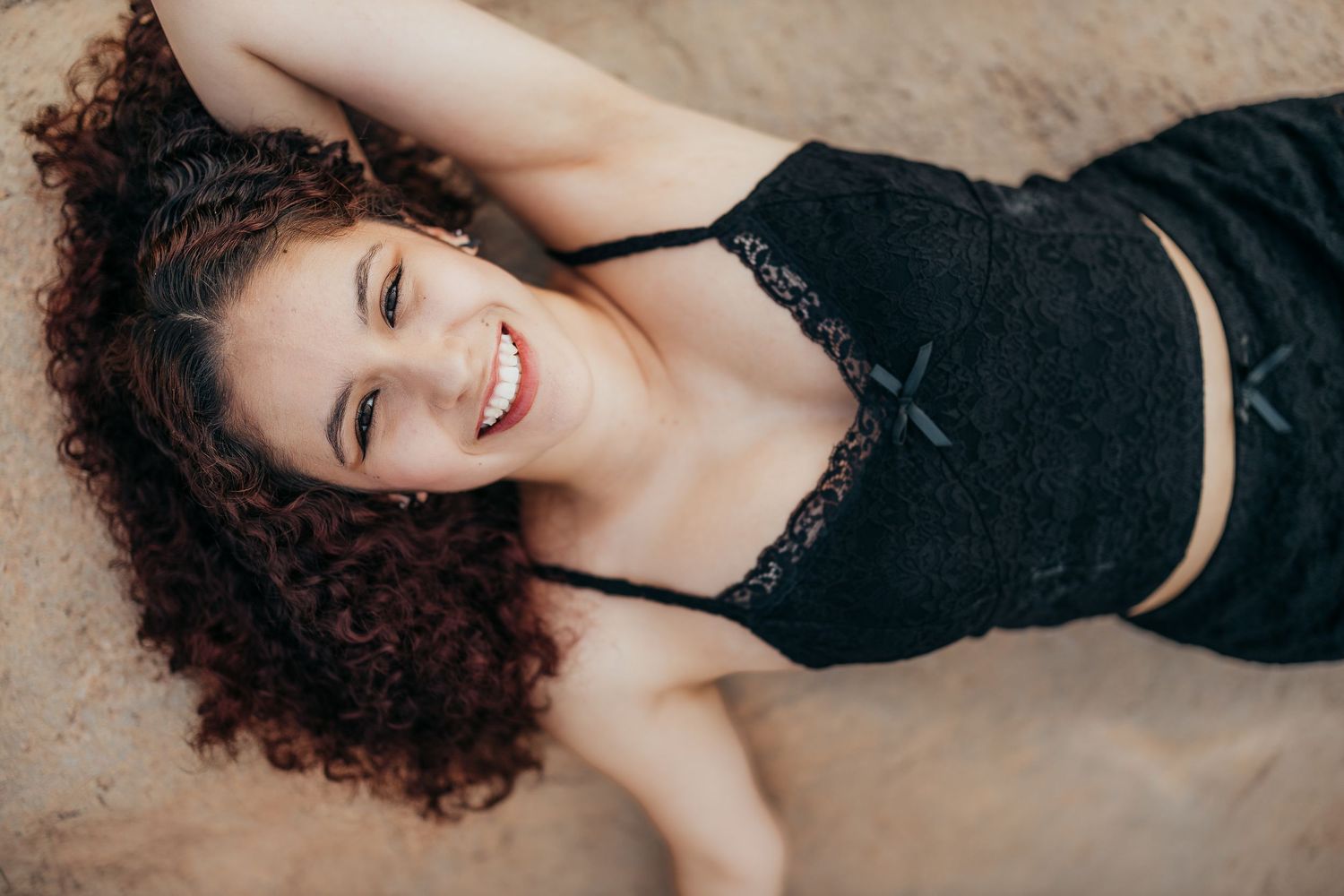 Person with curly hair wearing black outfit laying on sand during outdoor photo session.