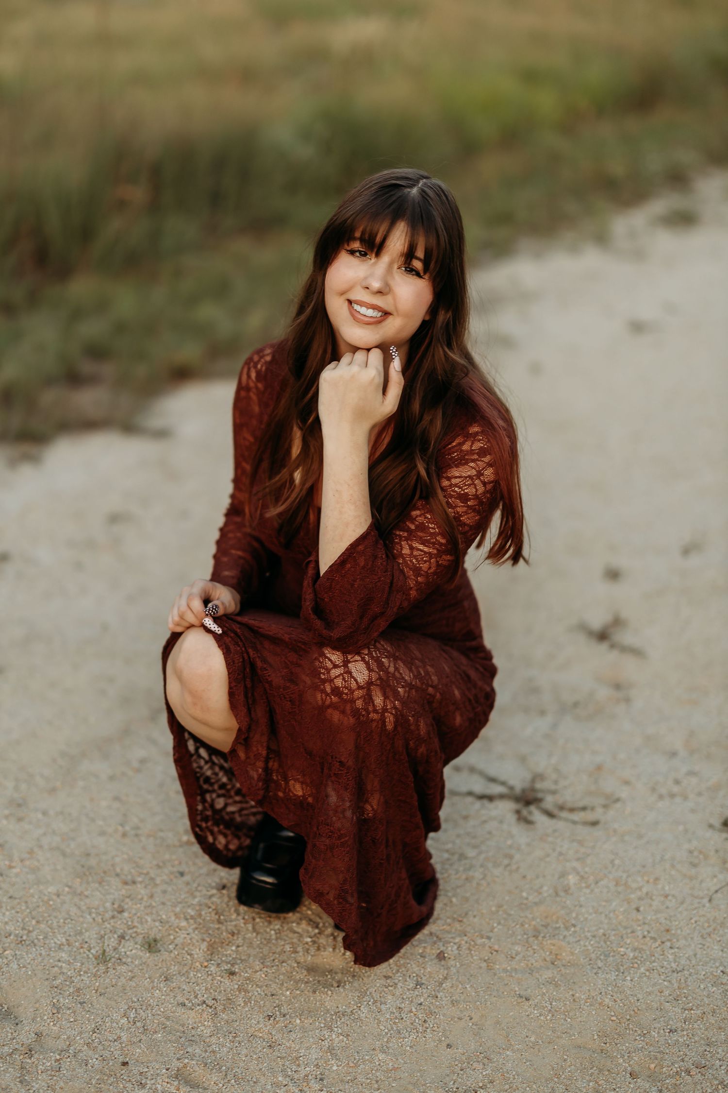 Woman in burgundy lace dress poses on sandy ground during outdoor photoshoot.