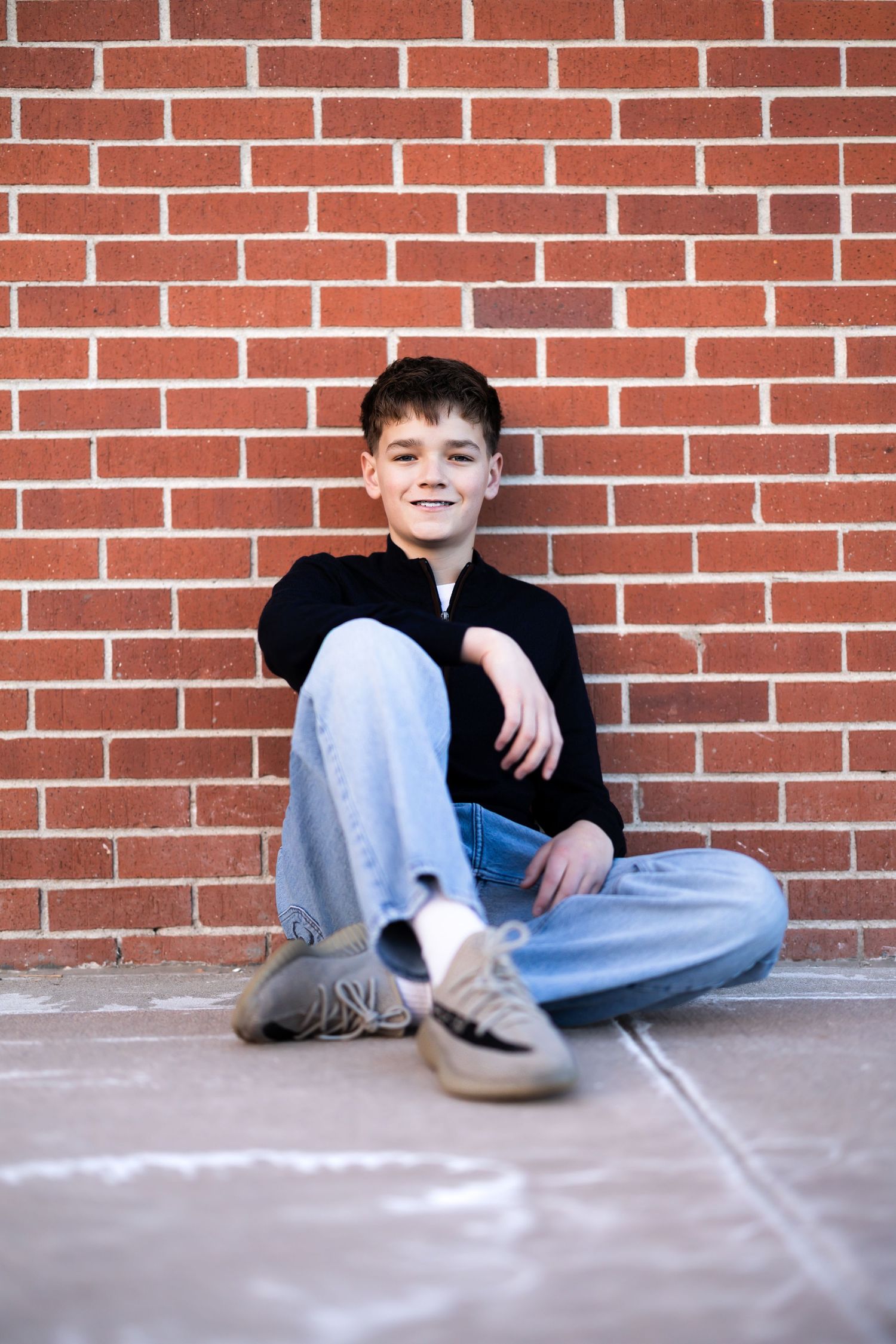 Person sitting against a brick wall wearing jeans and dark clothing in a casual pose.