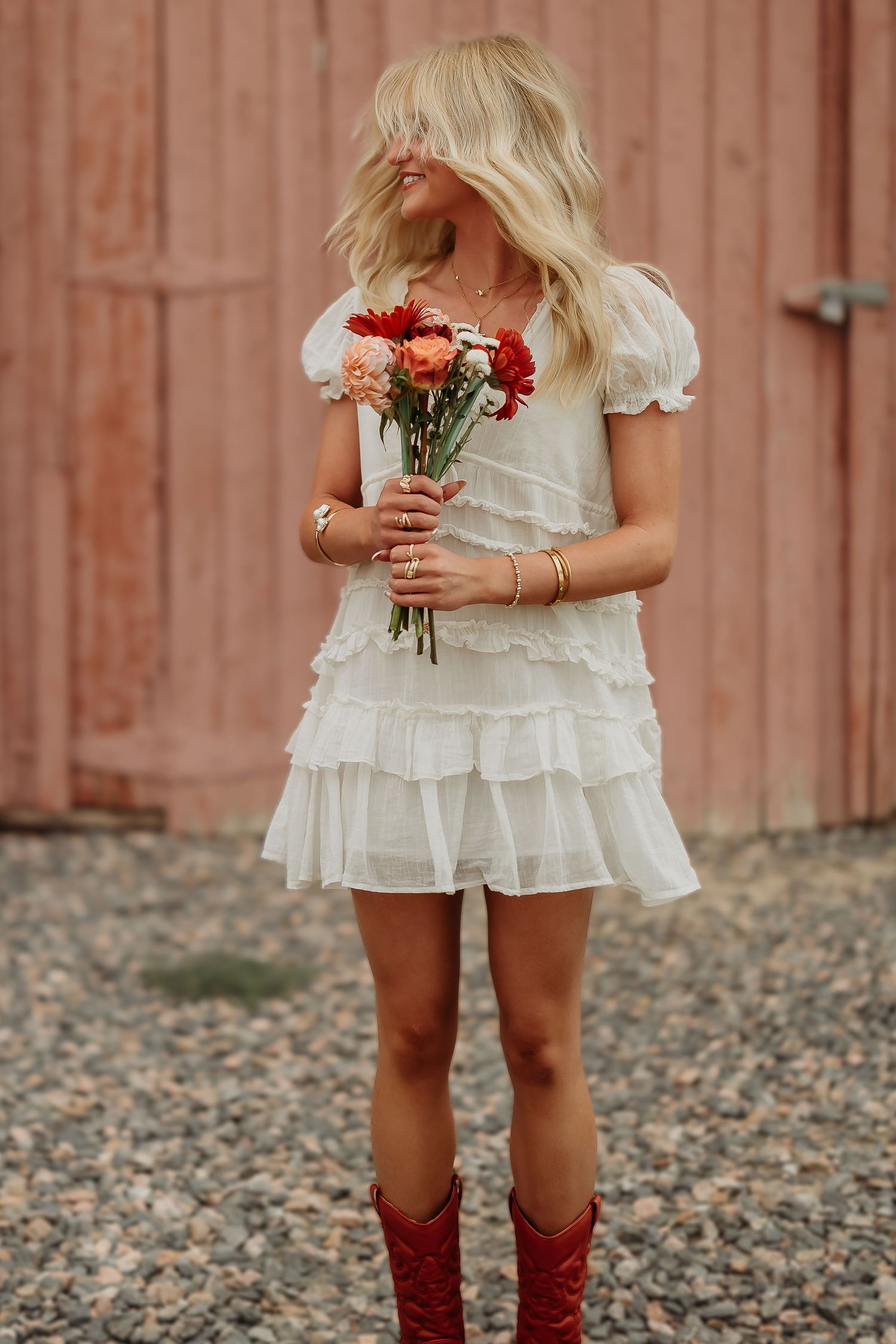 Person in white ruffled dress holding colorful flowers against rustic red barn background.