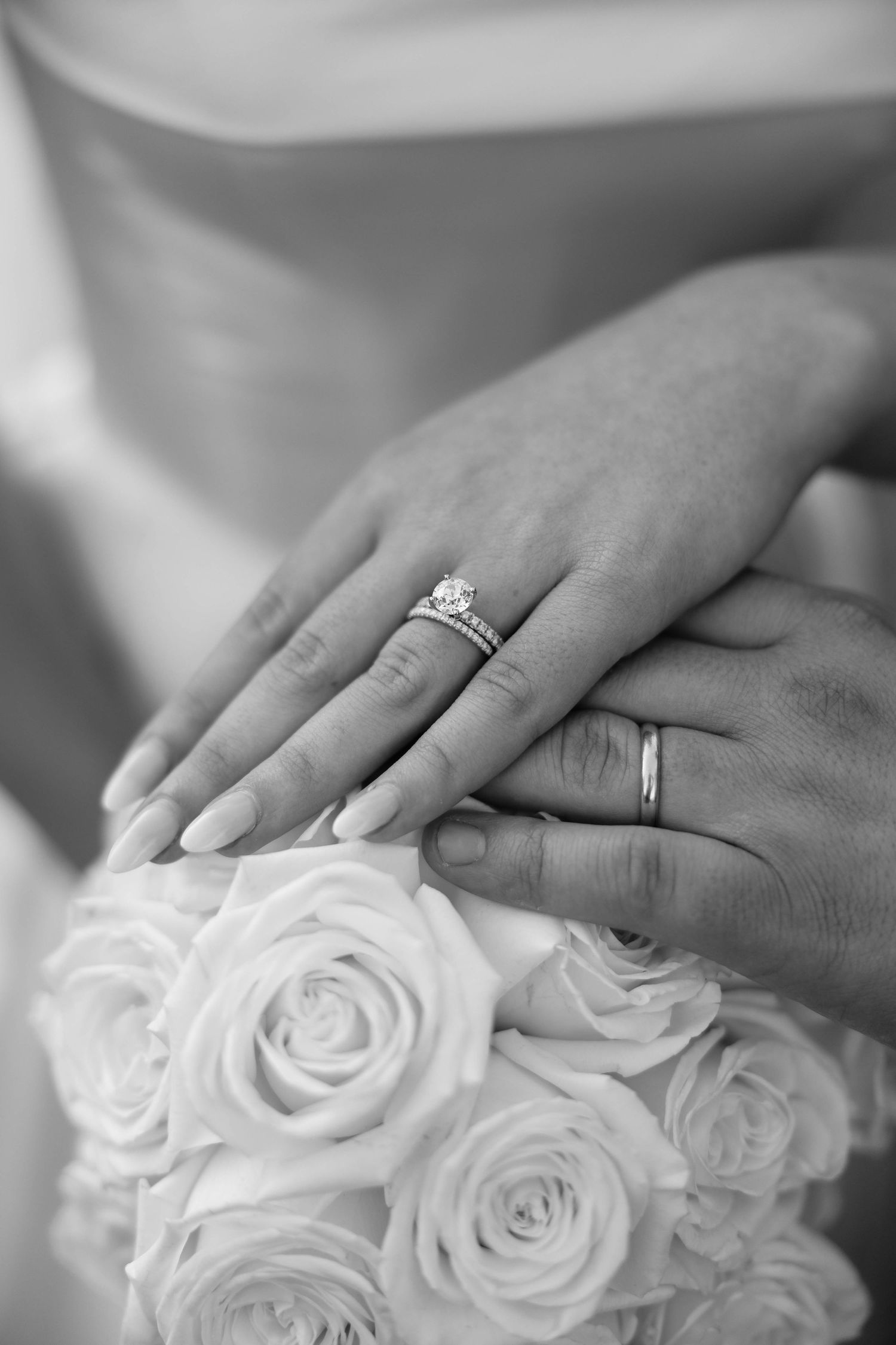 Black and white close-up of newlyweds hands with wedding rings resting on a rose bouquet.