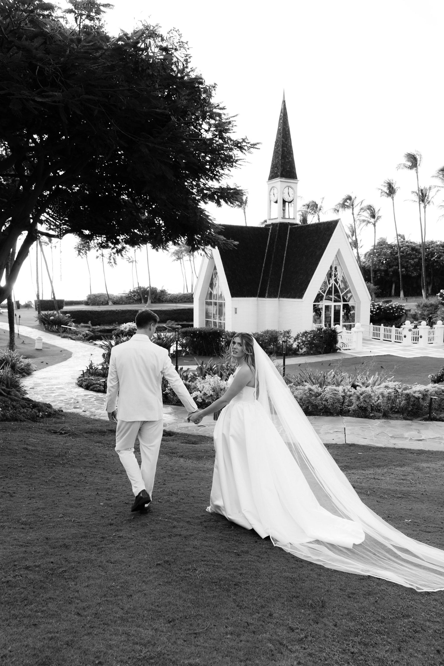 Black and white photo of bride and groom walking hand in hand toward a white chapel.
