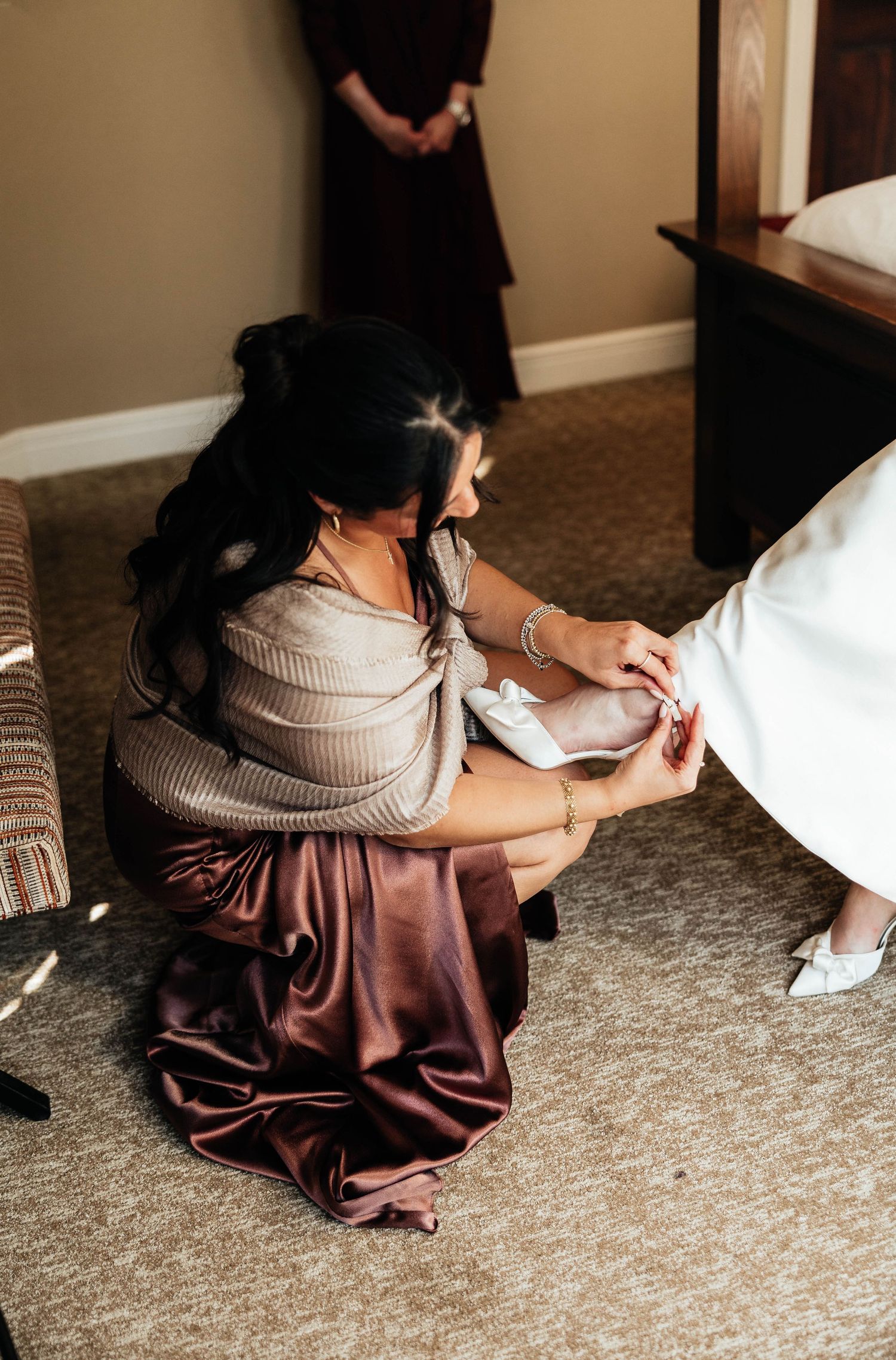 Bridesmaid kneels to help the bride put on her wedding shoes before the ceremony.