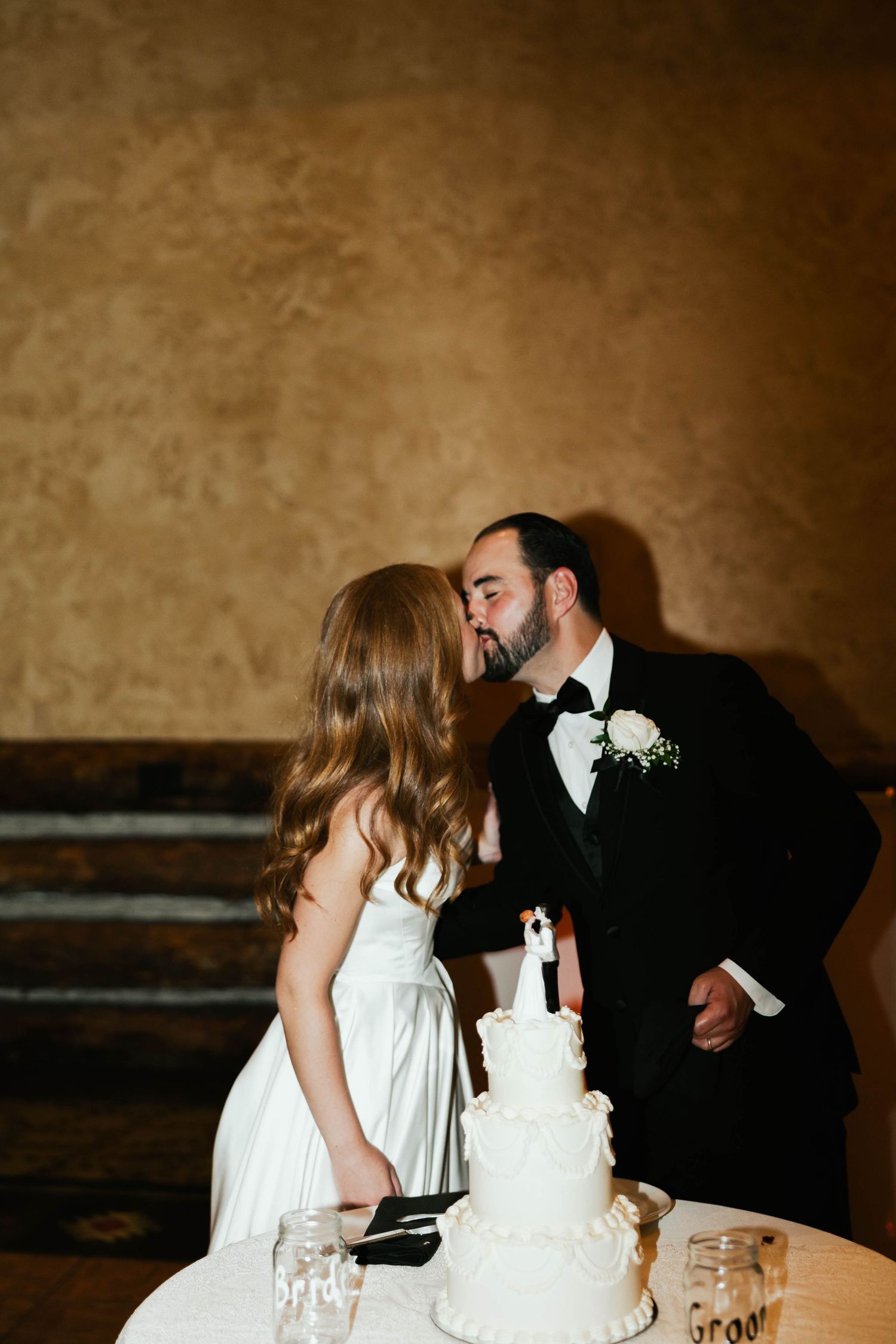 Bride and groom sharing a kiss behind their elegant white four-tier wedding cake at their reception.
