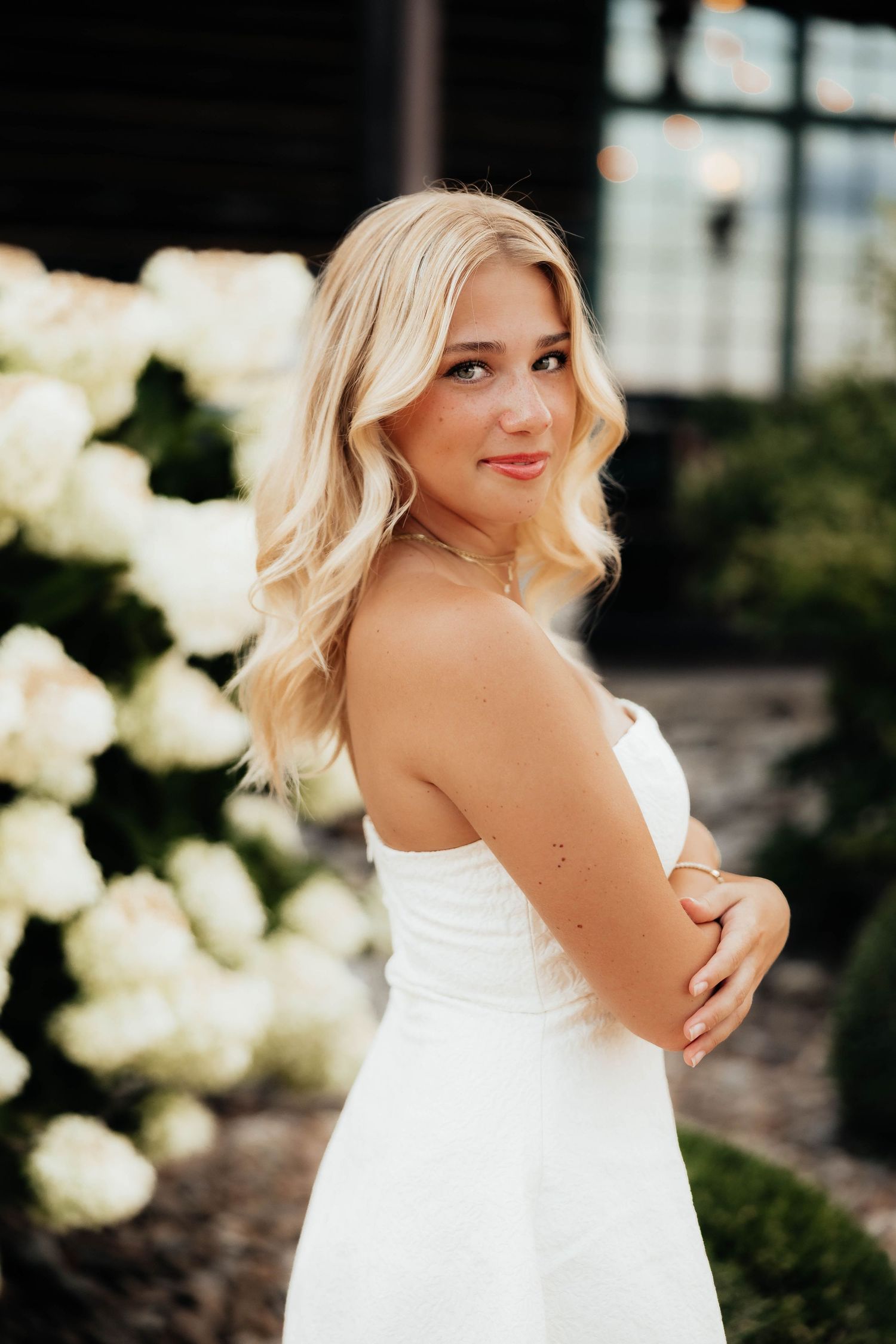 Smiling blonde woman in white strapless dress standing near white hydrangeas at outdoor venue.
