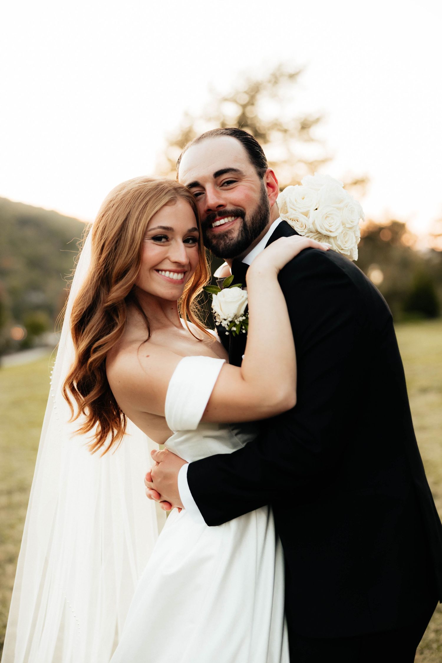 Joyful bride and groom embrace outdoors at sunset, groom holding white bouquet with rolling hills behind.