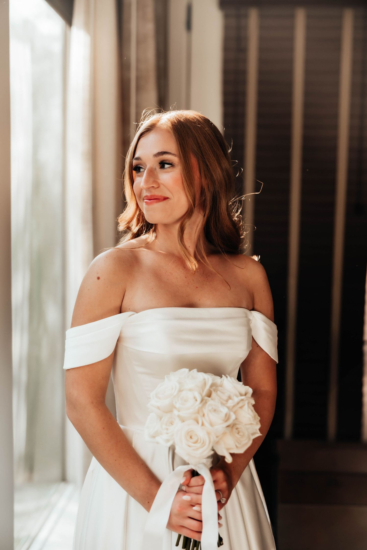 Bride in off-shoulder white satin gown holding white rose bouquet, gazing elegantly near a window in soft natural light.
