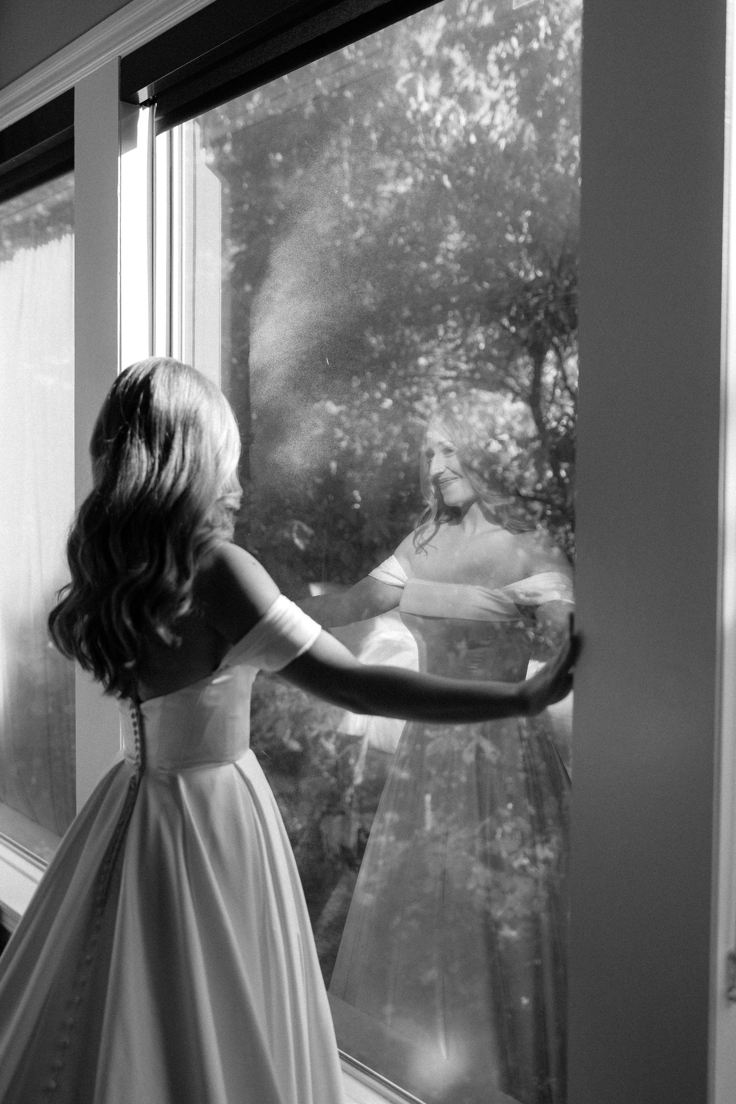 Black and white photo of bride in strapless gown with long hair, looking out a large window with tree reflections.