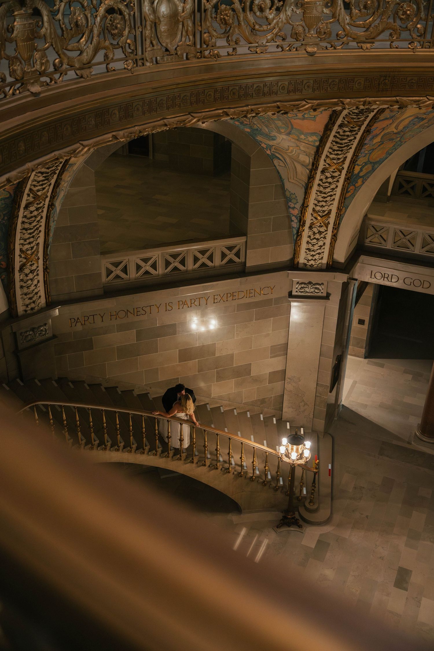 Bride and groom share romantic kiss on ornate grand staircase of historic building lit by candlelight.