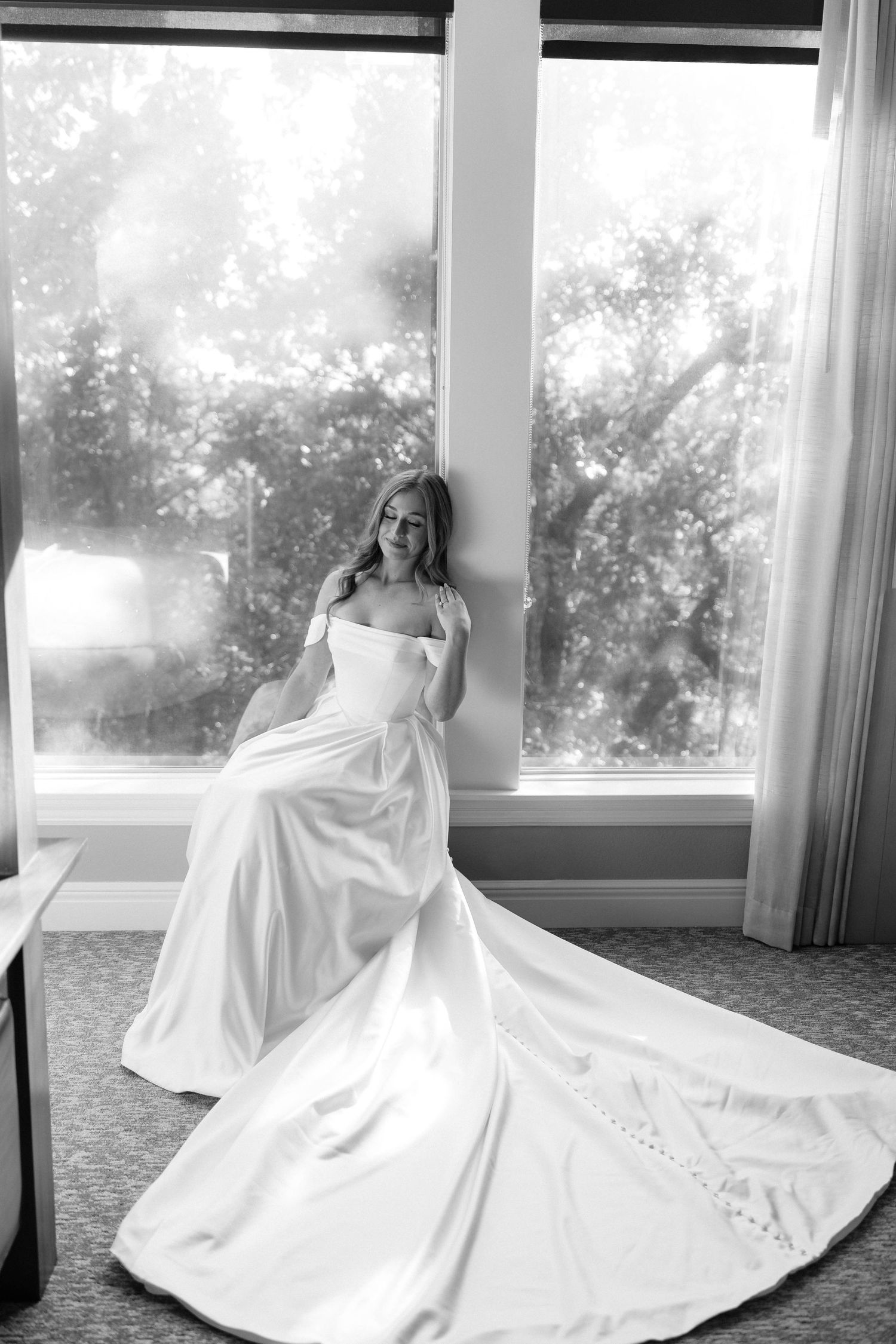 Black and white portrait of a bride in a dramatic gown with long train by a window.