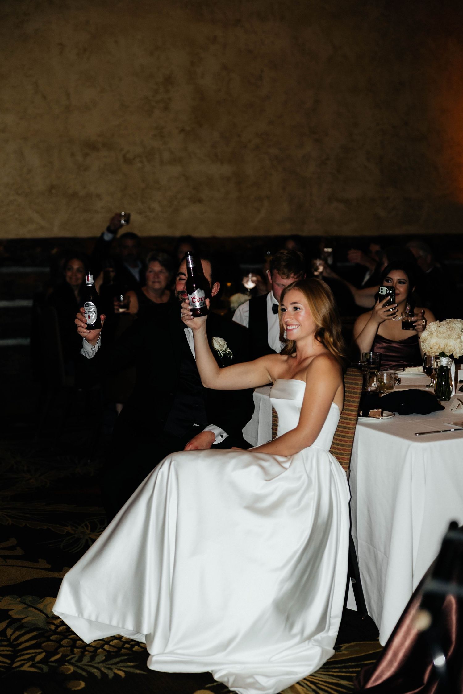 Smiling bride in white satin gown raising a bottle during a toast at her wedding reception.