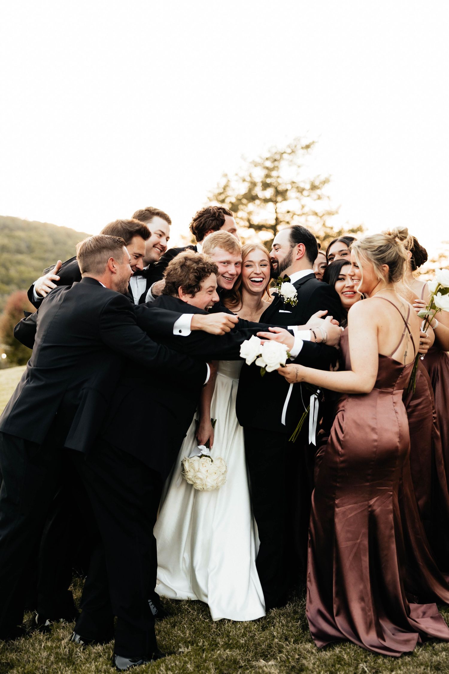 Bride and groom joyfully embraced by smiling wedding party outdoors in golden light.