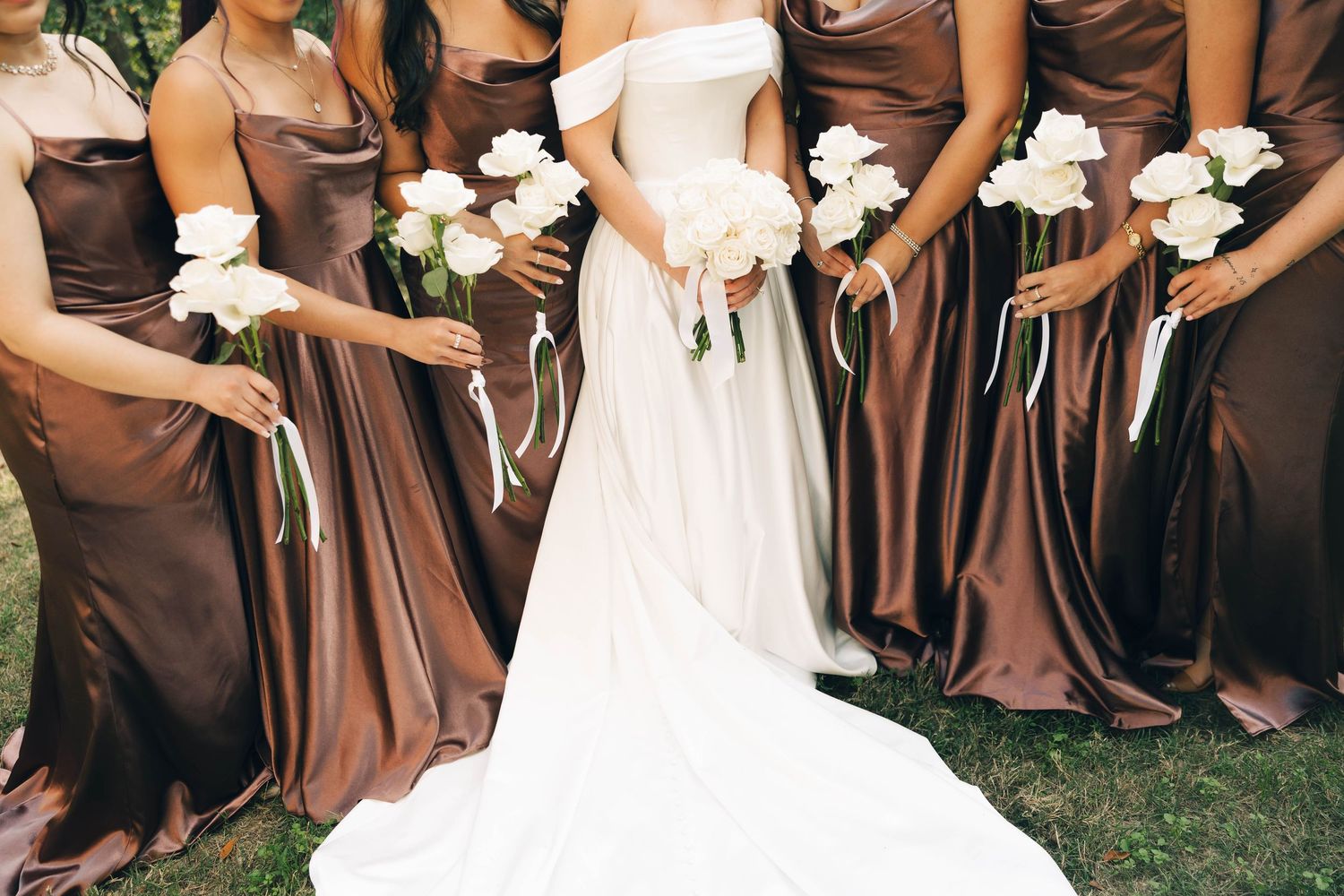 Close-up of bride in white gown flanked by bridesmaids in brown satin dresses holding white flowers.