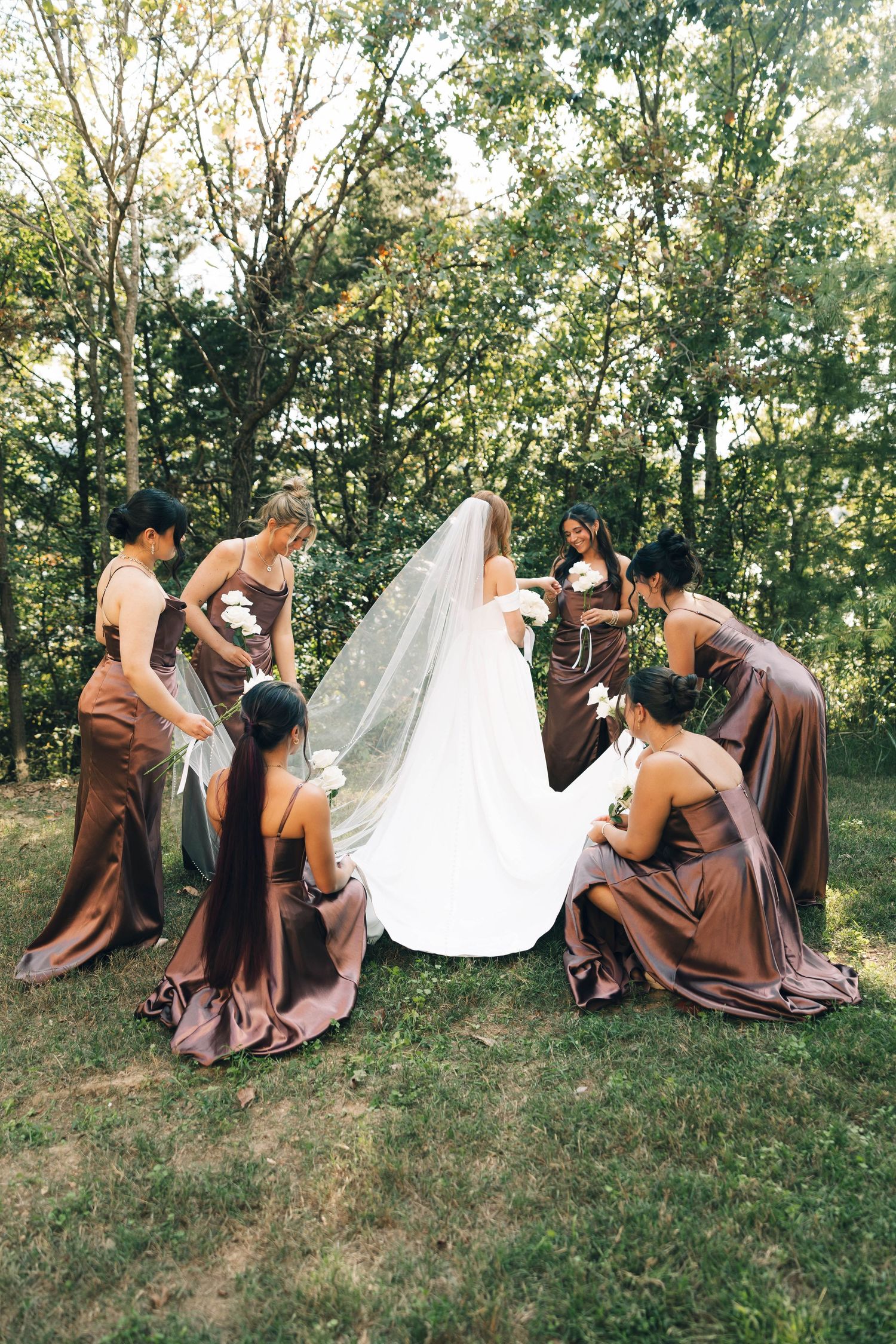 Bridesmaids in brown satin gowns kneel around bride adjusting her veil and train in forest setting.