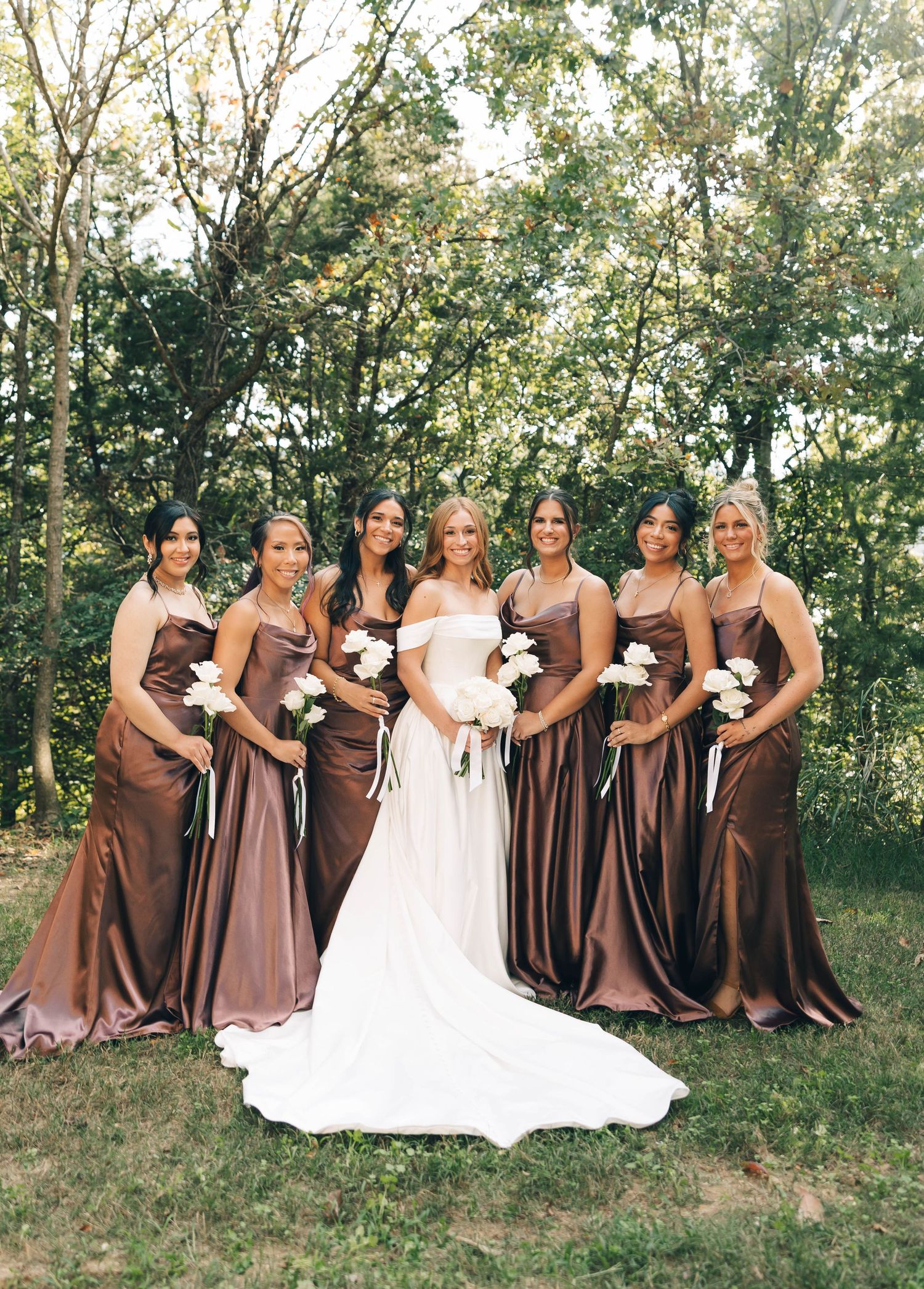 Bride in white gown poses with six bridesmaids in brown satin dresses holding white bouquets outdoors.