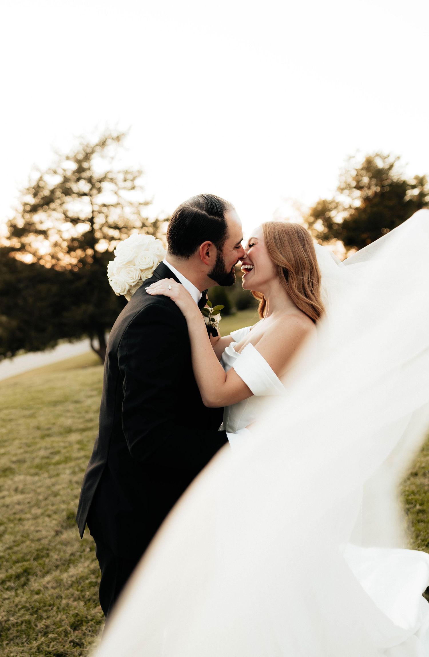 Laughing bride and groom nearly kiss outdoors at golden hour, veil flowing in the breeze.