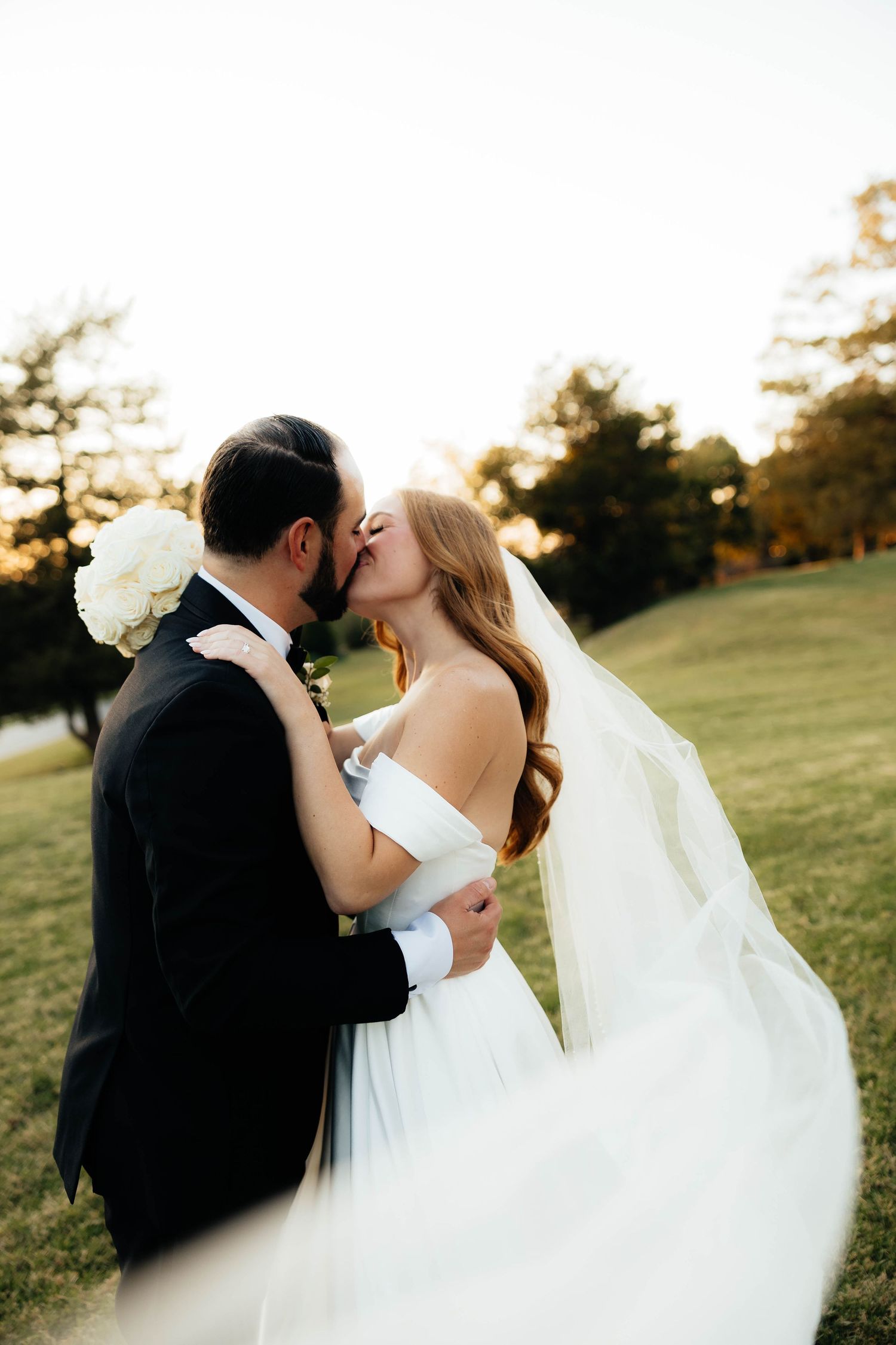 Newlyweds share a romantic kiss outdoors at sunset, bride in off-shoulder gown with flowing veil.