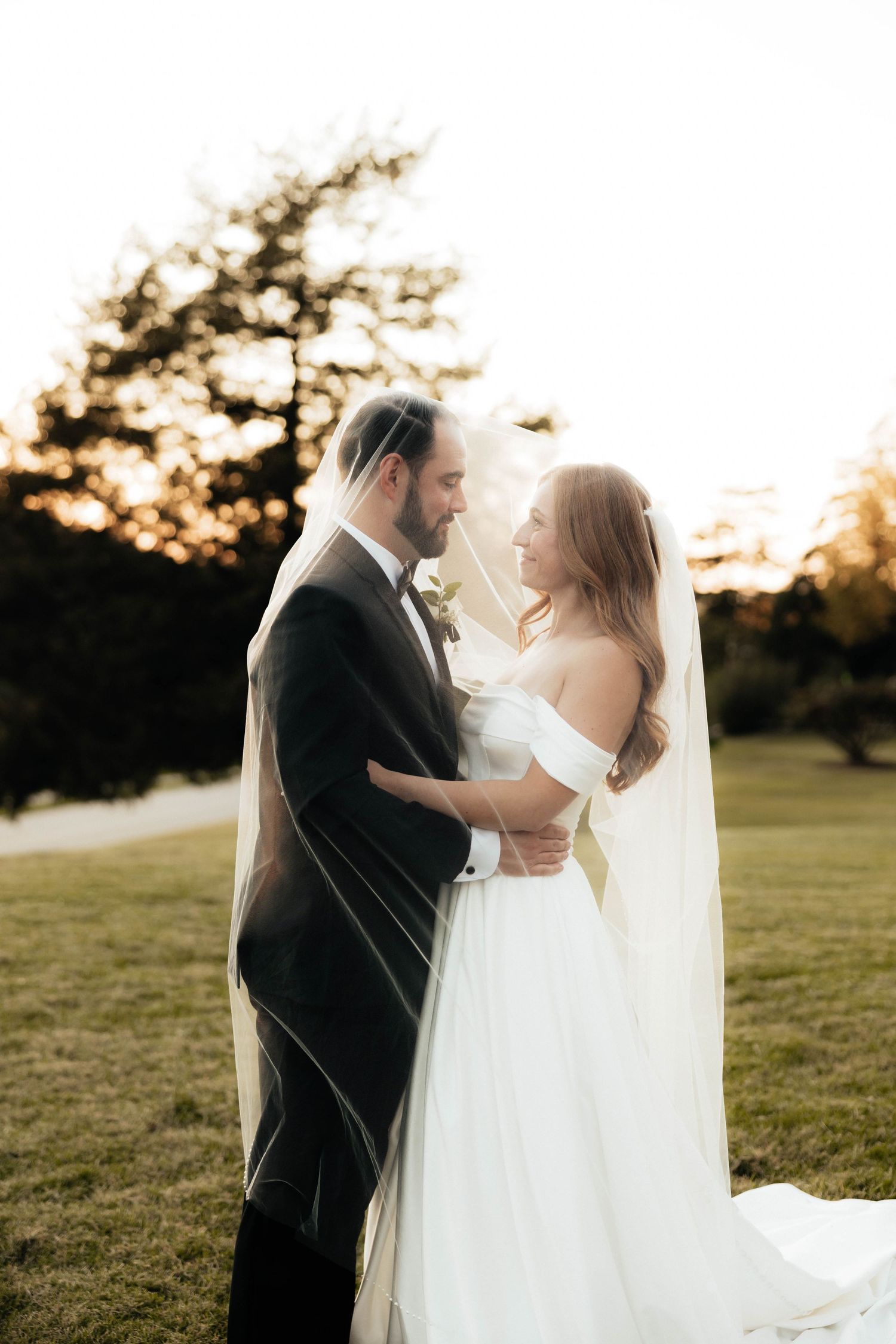 Bride and groom share an intimate moment under a flowing veil at golden hour in a lush green outdoor setting.