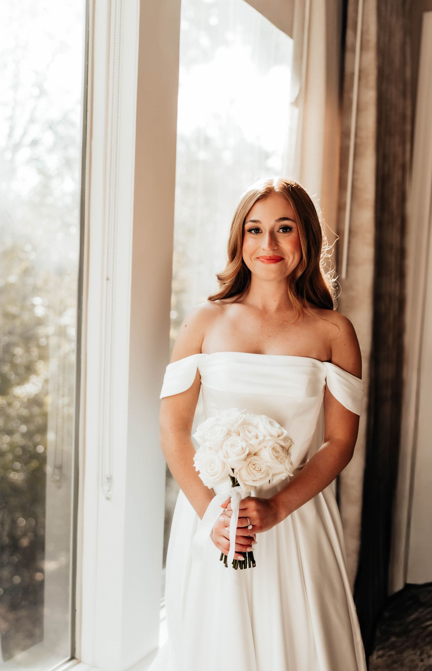 Elegant bride in off-shoulder white satin wedding dress holding white rose bouquet, standing by a bright window.