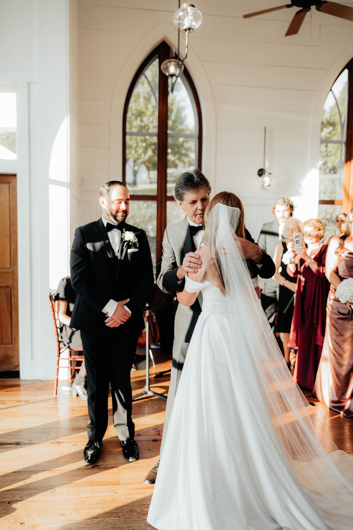 Father walking bride down the aisle toward smiling groom in a white chapel wedding.