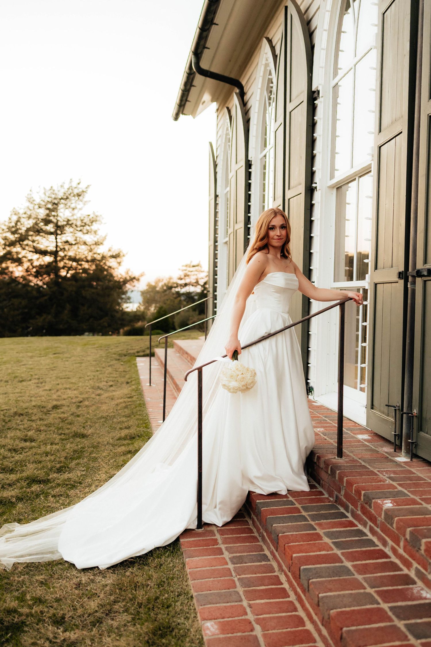 Bride in strapless white ball gown with long train holding bouquet, posing on brick steps of elegant venue at sunset.