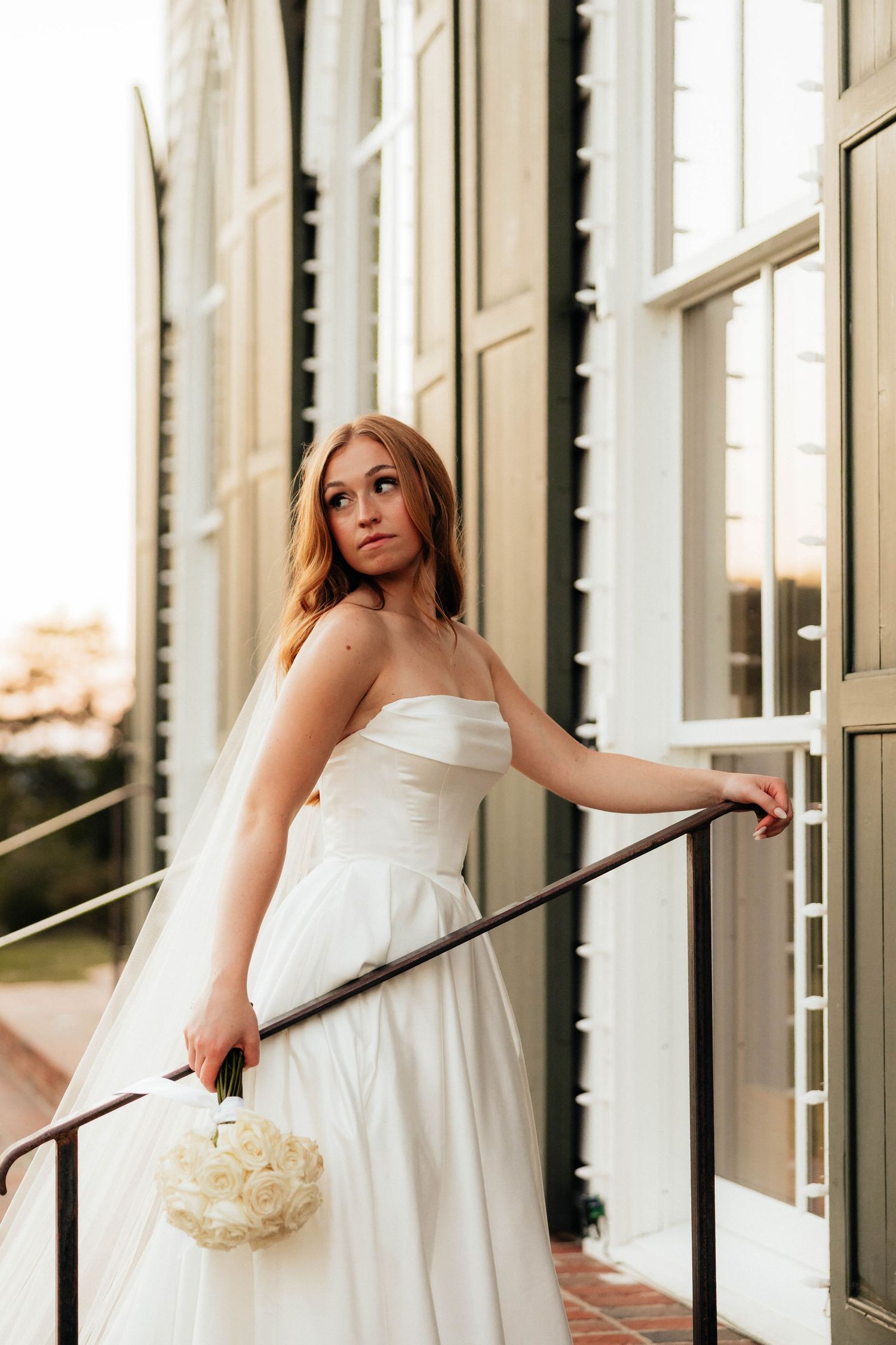 Bride in strapless white ball gown holding bouquet, leaning on railing outside elegant building at sunset.