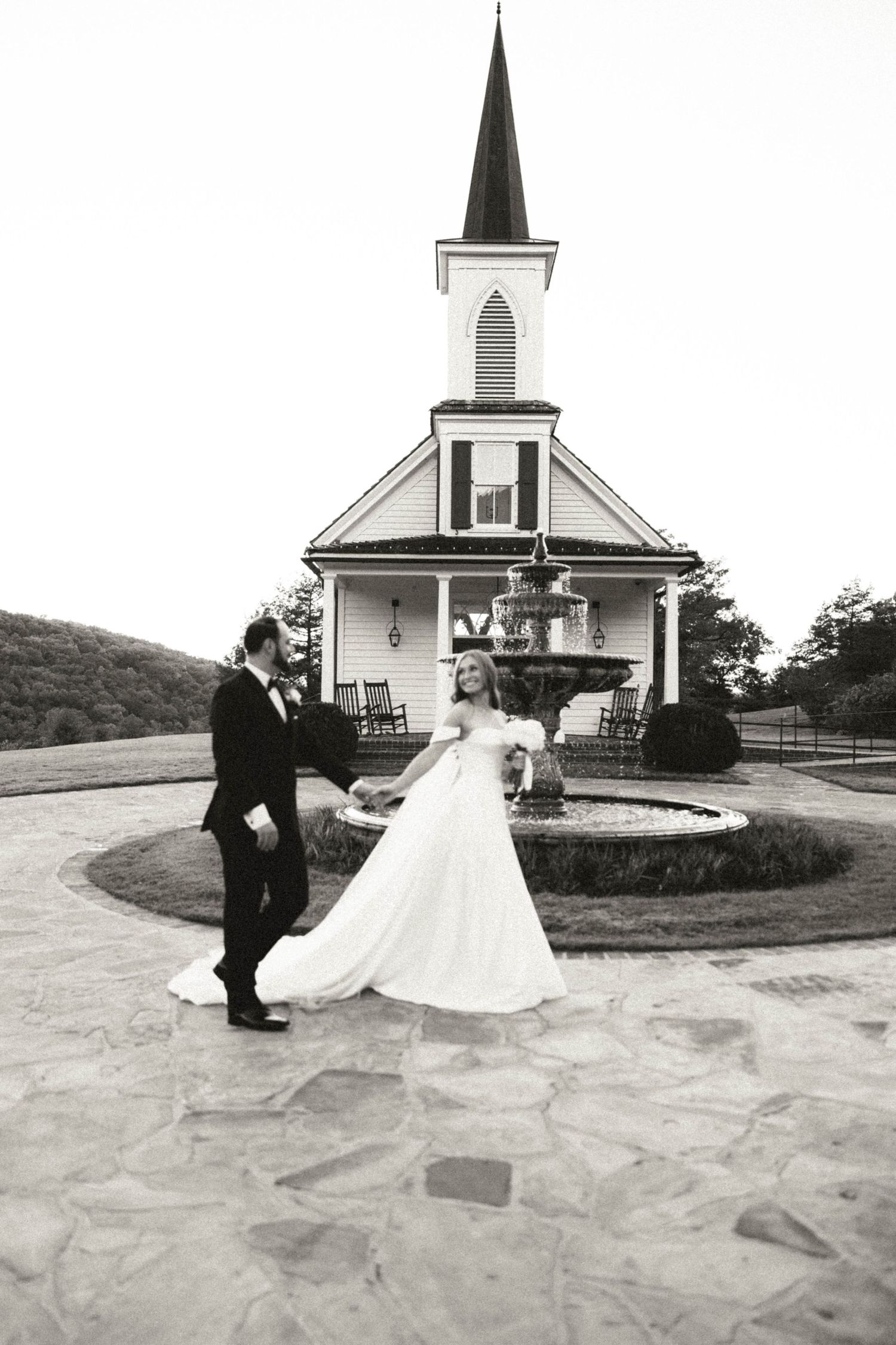 Black and white photo of bride and groom dancing in front of a white chapel with a steeple.