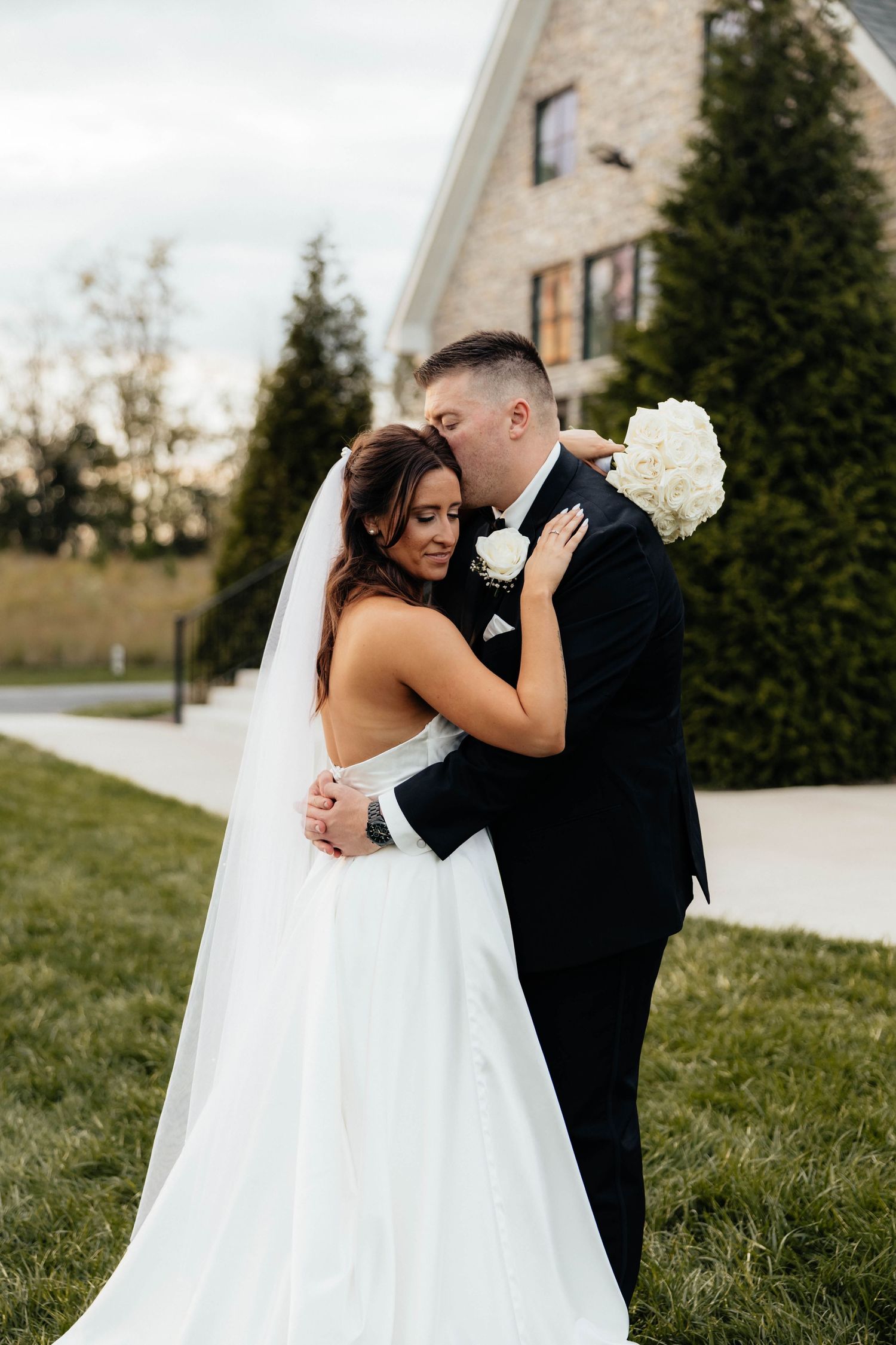 Groom in black tuxedo kisses bride in backless white gown outdoors near elegant stone building.