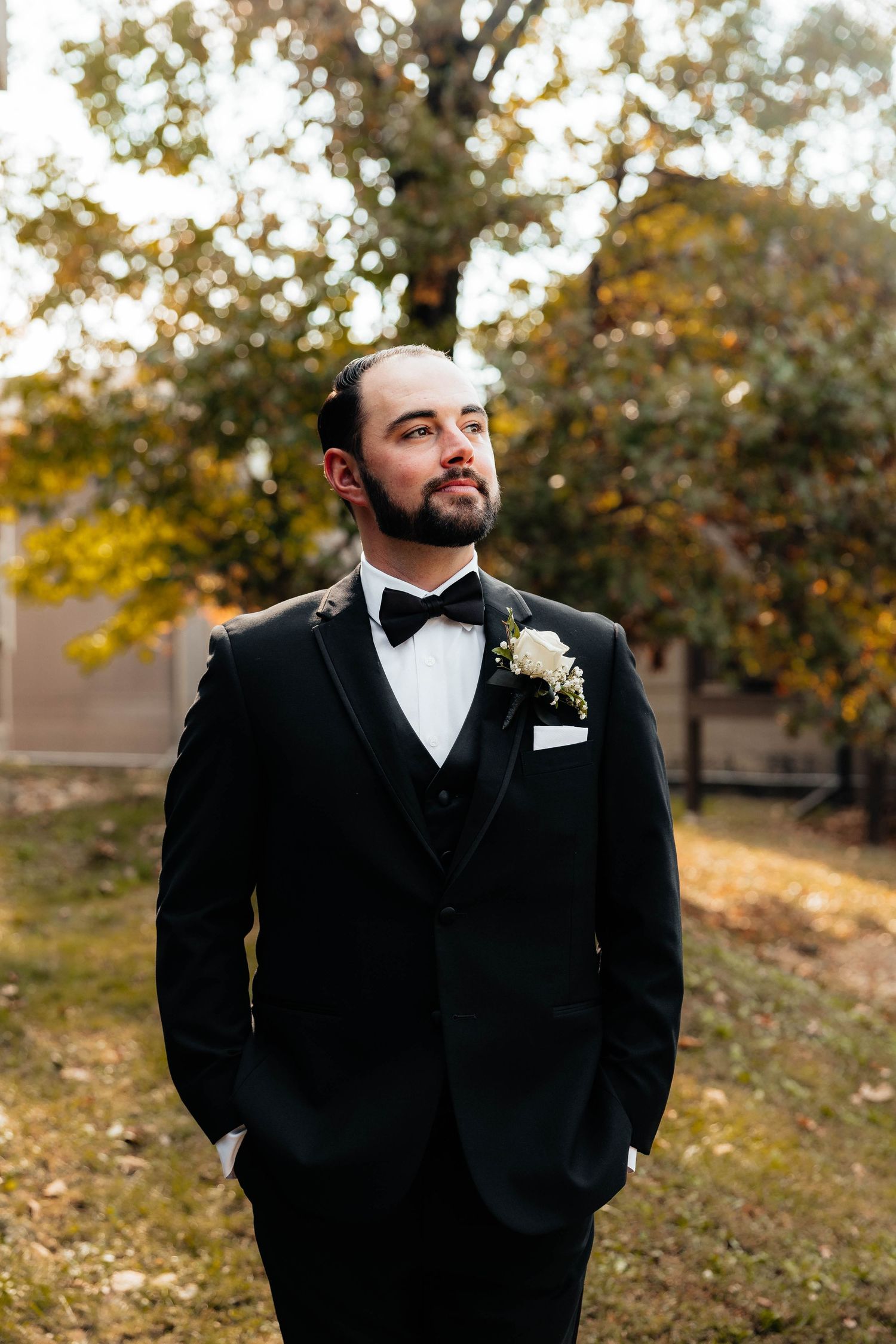 Groom in black tuxedo with bow tie and boutonniere standing outdoors among autumn trees.