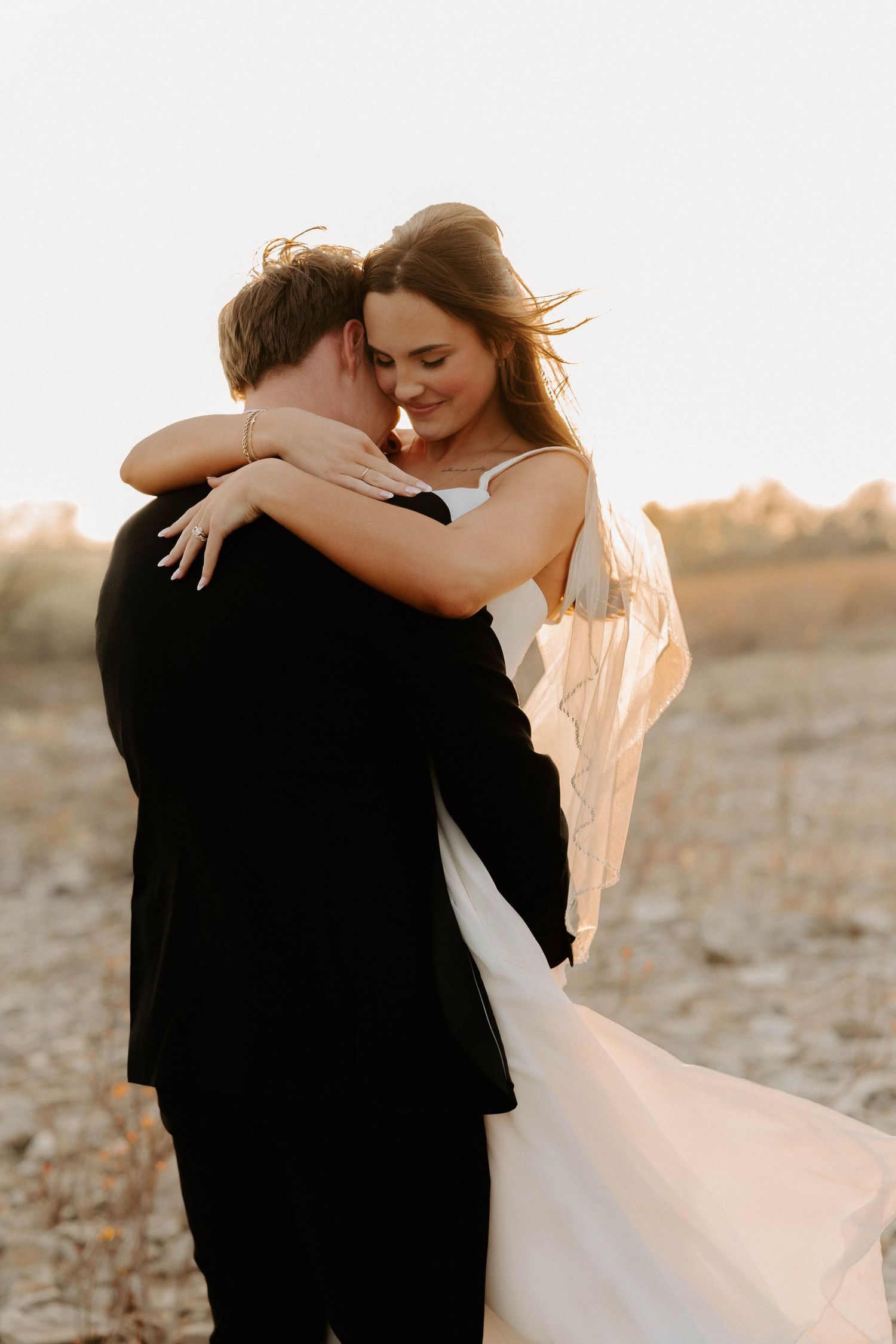 Bride and groom embracing in a golden sunlit field with the brides veil blowing in the wind.