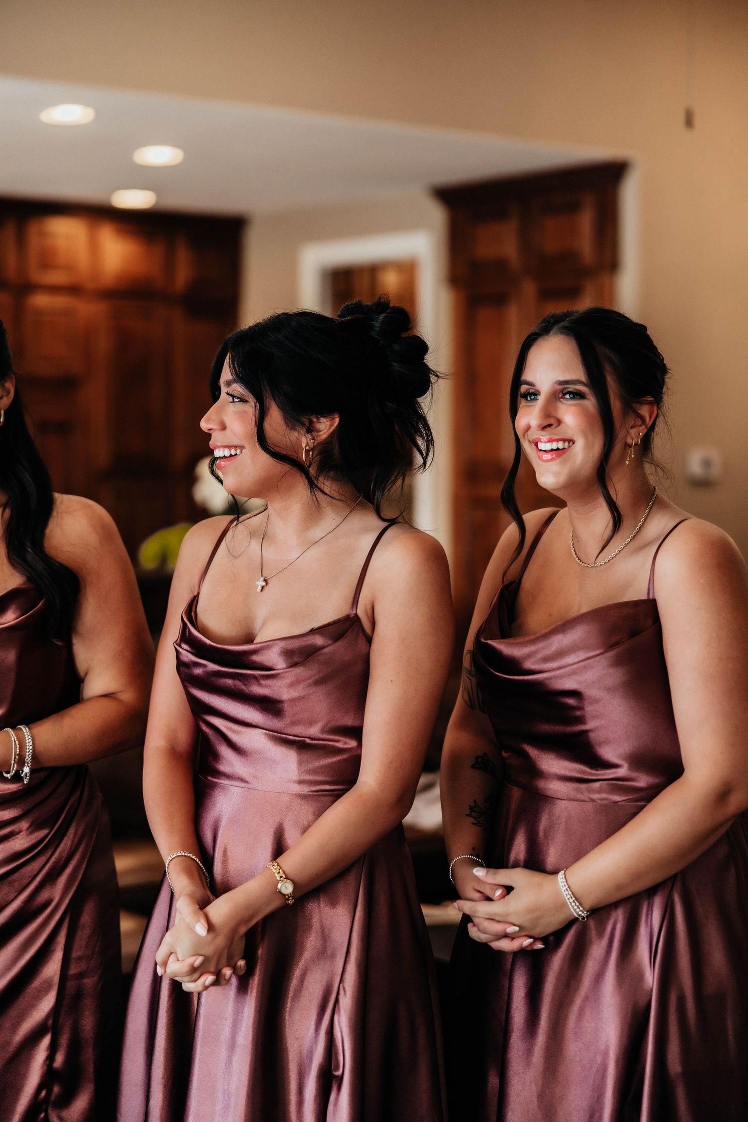 Smiling bridesmaids in dusty rose satin spaghetti-strap dresses standing together during wedding preparations indoors.