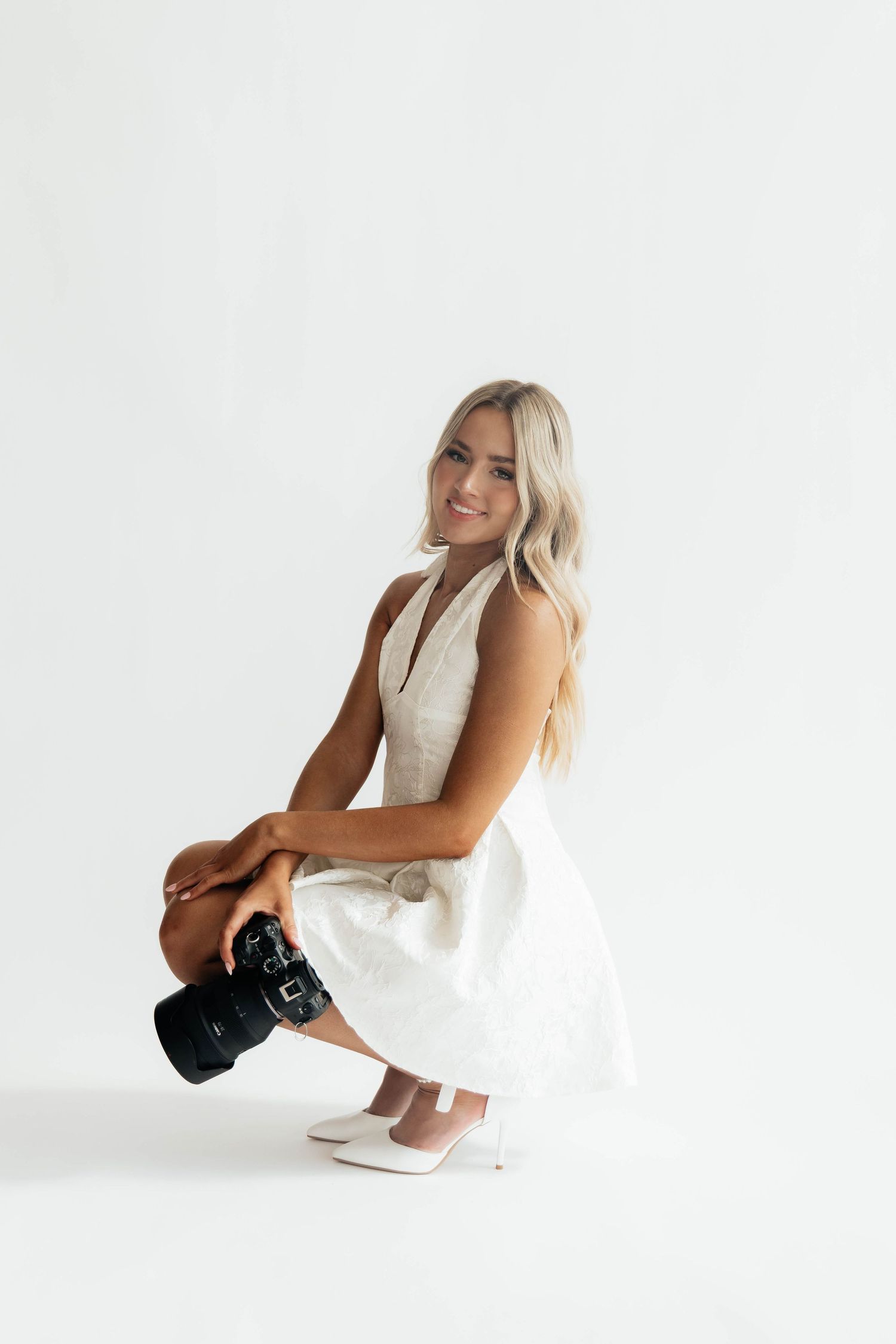 Smiling blonde woman in white dress crouching while holding a DSLR camera in a bright studio.