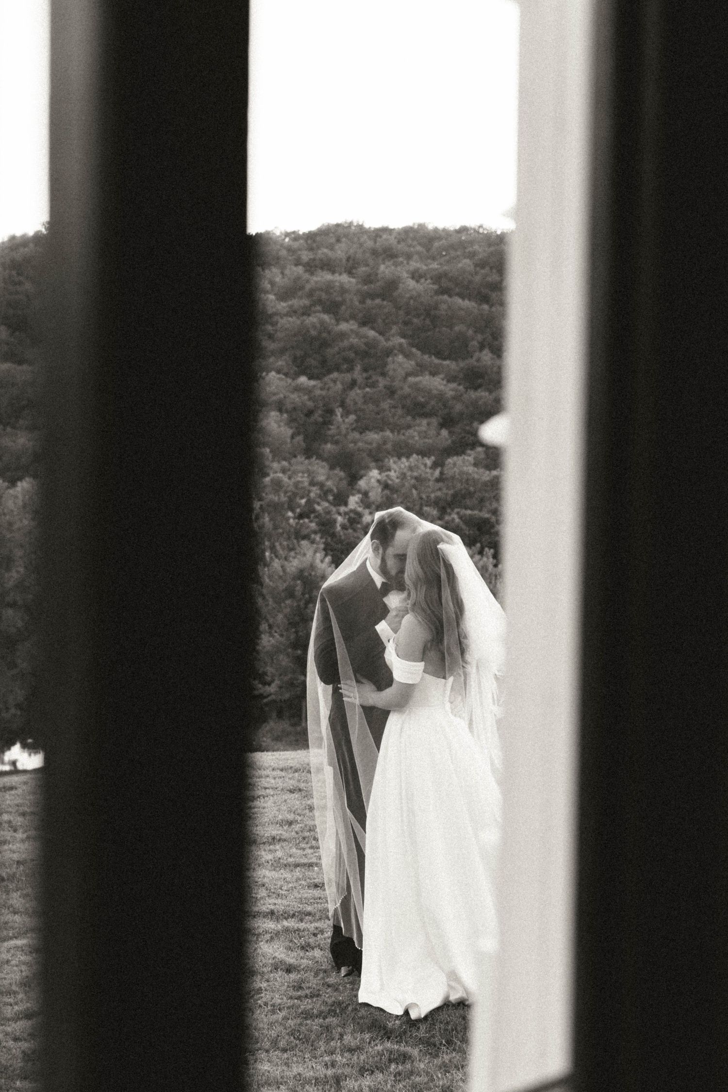 Black and white romantic photo of bride and groom kissing under veil framed by columns outdoors.