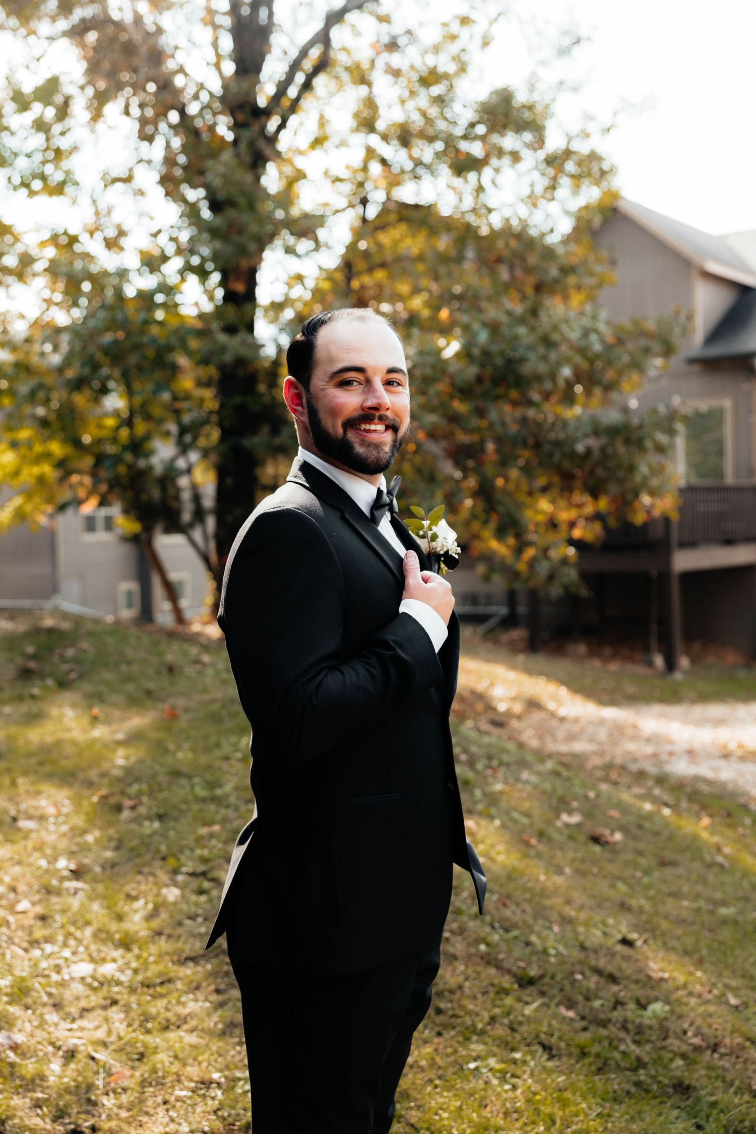 Handsome groom in black tuxedo with boutonniere smiling outdoors among autumn trees.