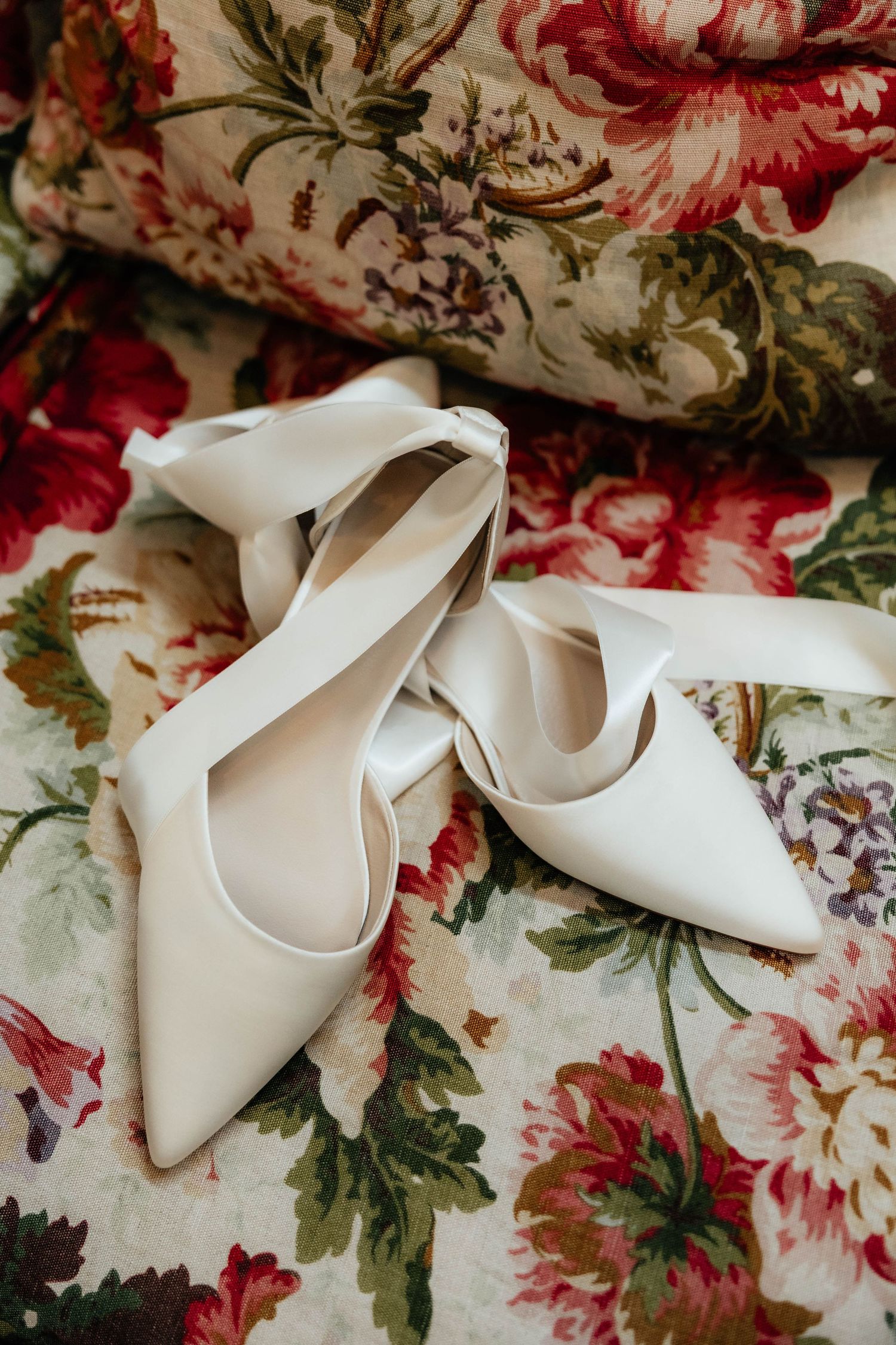 White pointed-toe bridal heels with ribbon ties resting on a floral patterned fabric.