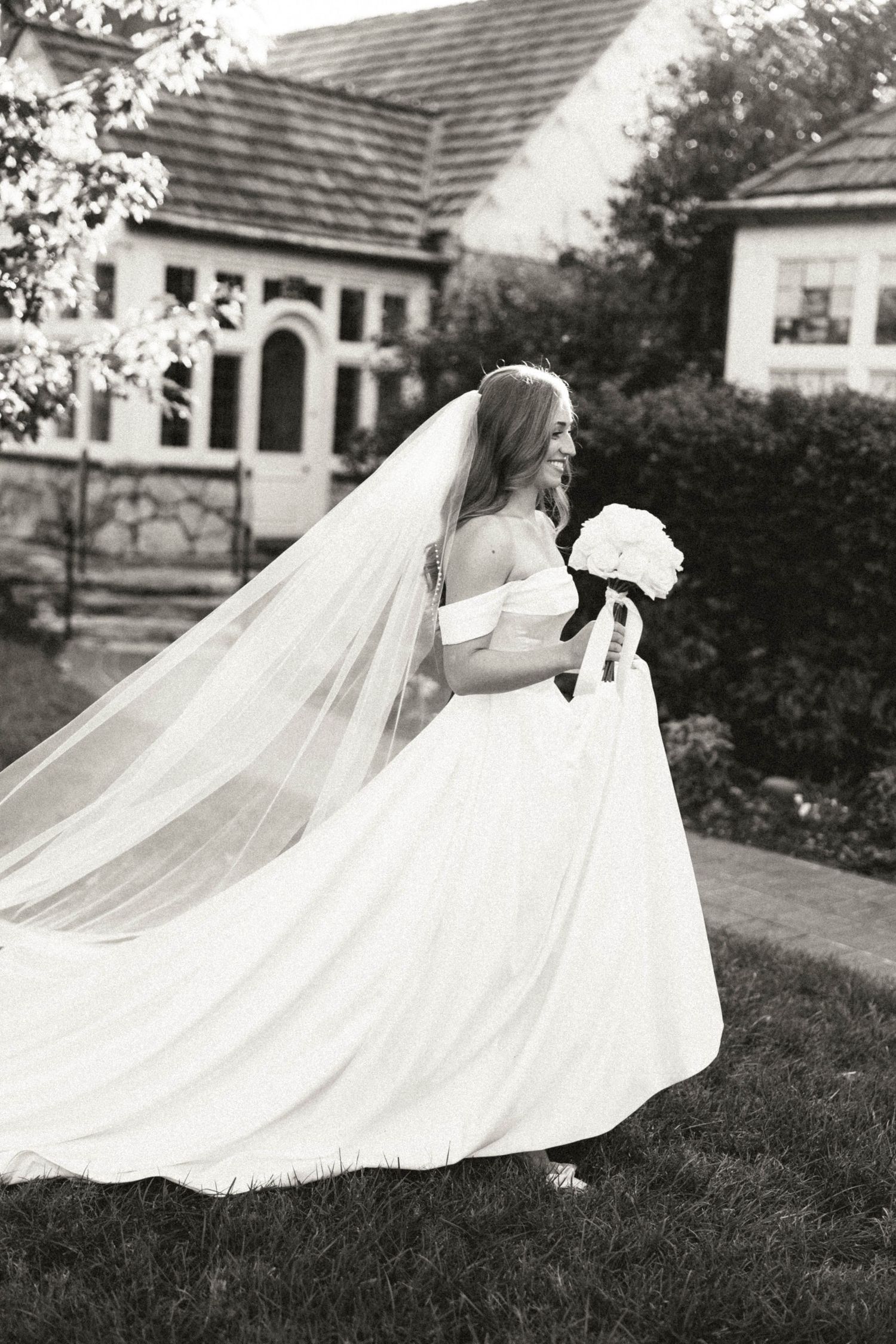 Black and white photo of joyful bride in off-shoulder gown with flowing cathedral veil walking outdoors.