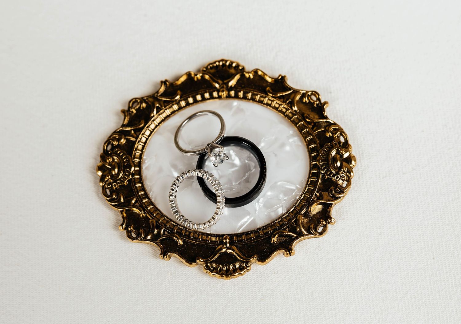 Gold ornate oval frame displaying wedding rings and jewelry on white background.