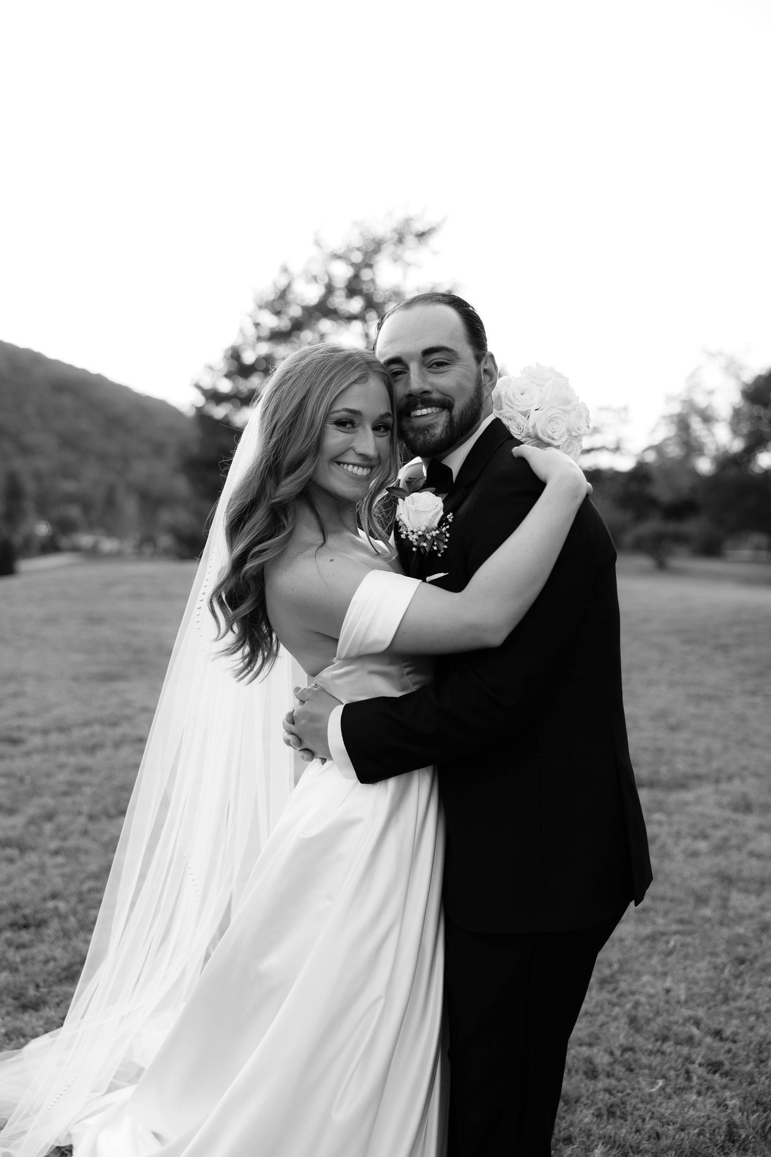 Black and white portrait of smiling bride and groom embracing in a scenic outdoor setting with mountains.