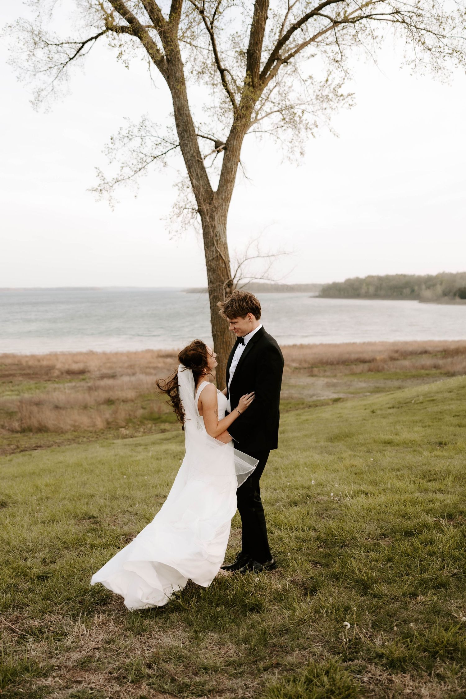 Bride and groom sharing a tender moment under a bare tree near a serene lakeside.